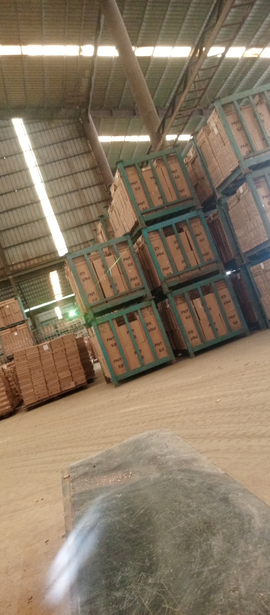 The image depicts an indoor warehouse scene in Magboro, Nigeria. Stacks of cardboard boxes, some packed in green metal cages, are scattered throughout the warehouse. A large metal support beam runs across the ceiling. The floor appears to be concrete. The lighting is provided by sunlight coming through the roof and some electric lights. There are no people visible in the frame.