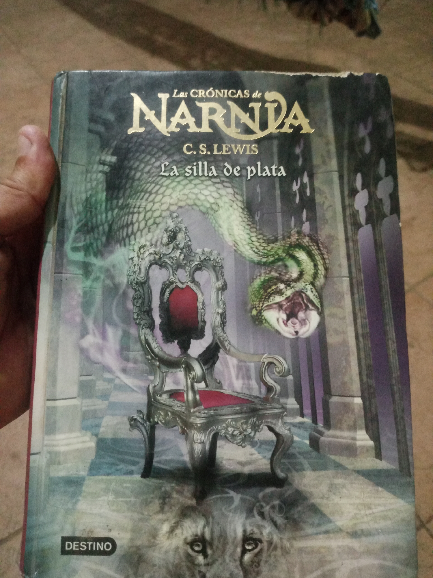 A book cover is held by a person's hand. The book is titled "Las Crónicas de Narnia: La Silla de Plata" by C.S. Lewis. The cover features an illustration of a green serpent with an open mouth, a detailed ornate chair, and the face of a lion. The background depicts a hall with columns and a patterned floor. The book cover has signs of wear and tear, particularly along the edges. The background environment is out of focus and appears to be a natural setting. No specific location in Santa Ana, Venezuela, can be identified from the image.