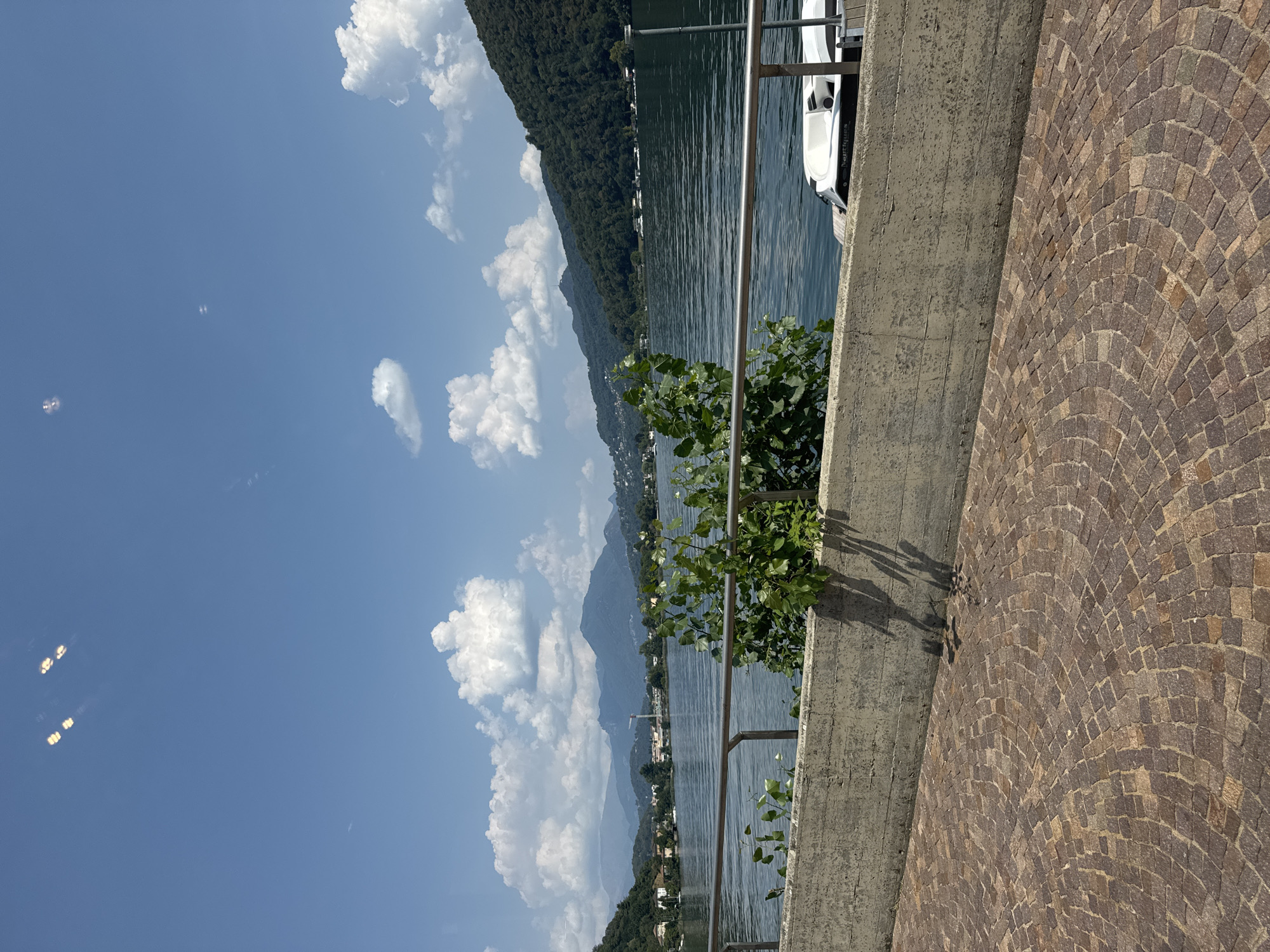The image captures a serene waterside scene in Lavena Ponte Tresa, Italy. In the foreground, a textured cobblestone patio is visible, with a weathered concrete barrier running along its edge. A metal railing extends across the barrier, partially obscured by lush green foliage. Beyond the railing, a tranquil body of water reflects the clear blue sky dotted with fluffy white clouds. On the far side of the water, rolling green hills rise, dotted with buildings and trees, creating a picturesque backdrop. A white boat is moored to a wooden dock on the right side of the water. The overall atmosphere is peaceful and idyllic, typical of a lakeside setting.