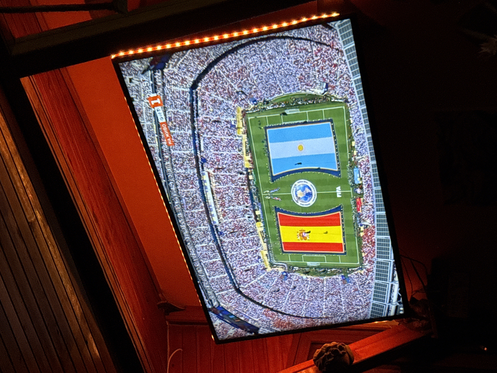 The image shows a television screen displaying a soccer match between Spain and Argentina in a packed stadium. The flags of both countries are visible on the field, along with the FIFA logo. The screen is mounted on a wall or structure that is lit with red and orange lights, creating a warm and festive atmosphere. The surrounding environment appears to be an outdoor setting, possibly a patio or covered area, with wooden beams and red-painted surfaces. The overall impression is one of a sports viewing party or event.
The city of Mineralni Bani, Bulgaria is not depicted or suggested in this image.