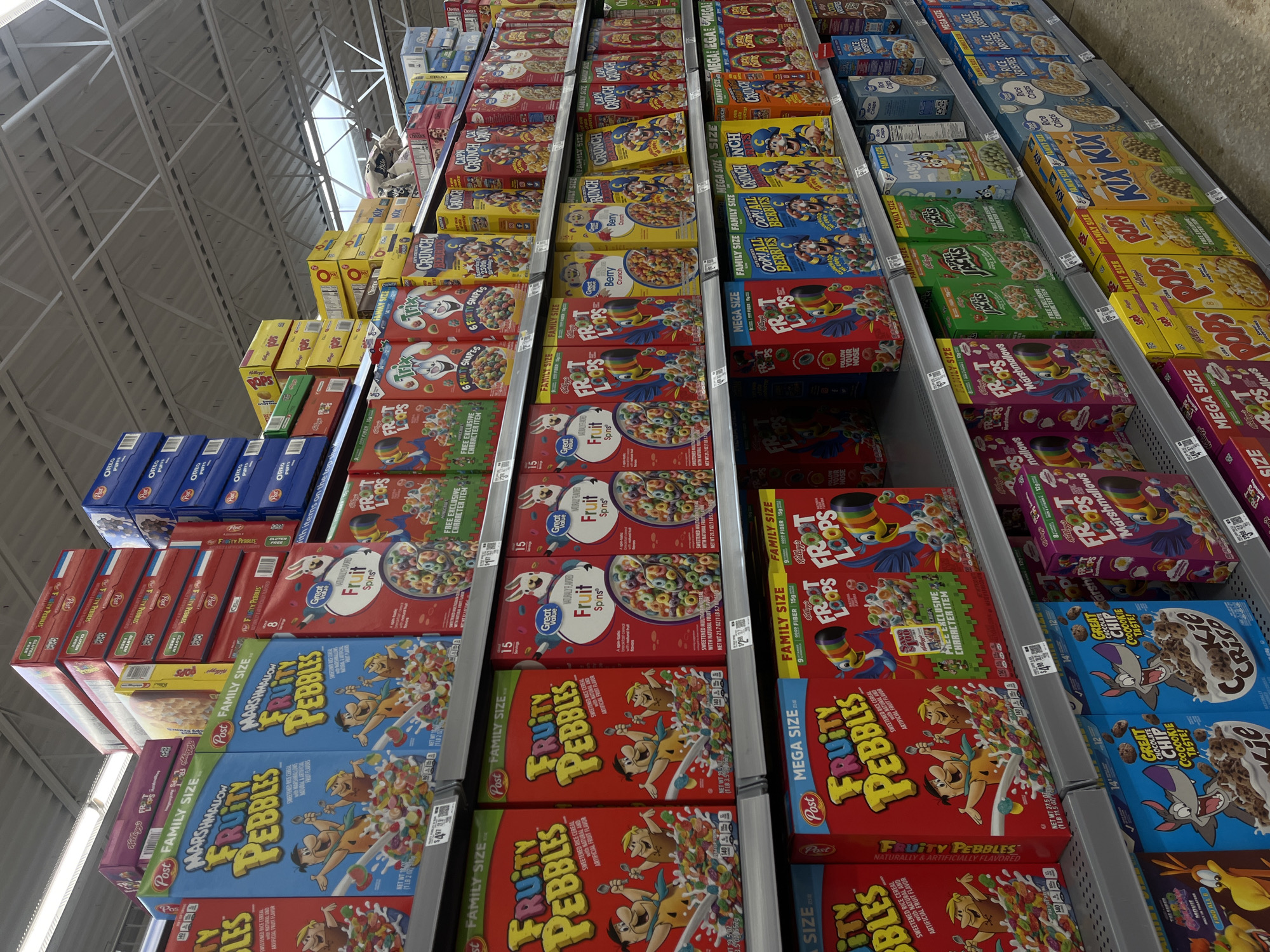 The image shows shelves stocked with various breakfast cereals, primarily Fruit Pebbles, Fruity Loops, and Cap'n Crunch. The shelves are arranged vertically, with multiple rows of cereal boxes stacked on each shelf. The packaging varies, with "Family Size" and "Mega Size" options visible. Other cereal brands like Kix and Rice Krispies are also discernible. The setting appears to be a supermarket aisle due to the systematic shelving and the variety of packaged goods. The lighting suggests an indoor environment with artificial illumination. There are no people visible in the shot. The location is likely a grocery store in San Antonio, United States of America.
