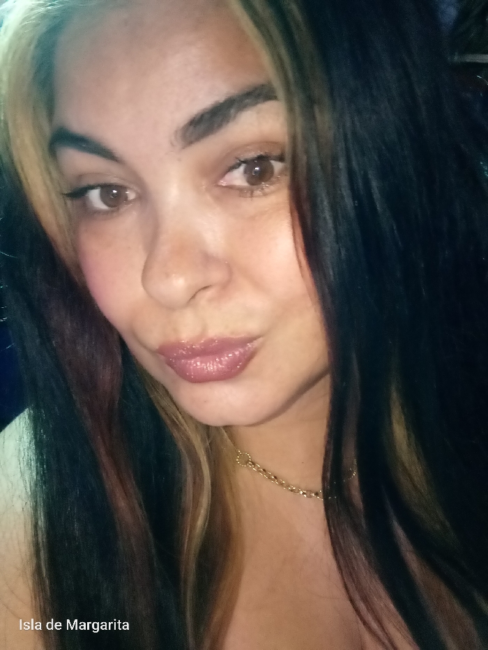 A woman's selfie shows her face closely. Her hair is long, dark, with blonde and red streaks. She has brown eyes, dark eyebrows, and full, glossy lips. She is wearing a delicate gold chain necklace. The photo has a slightly blurry quality, and the lighting is somewhat dim. The text "Isla de Margarita" is written at the bottom of the picture.