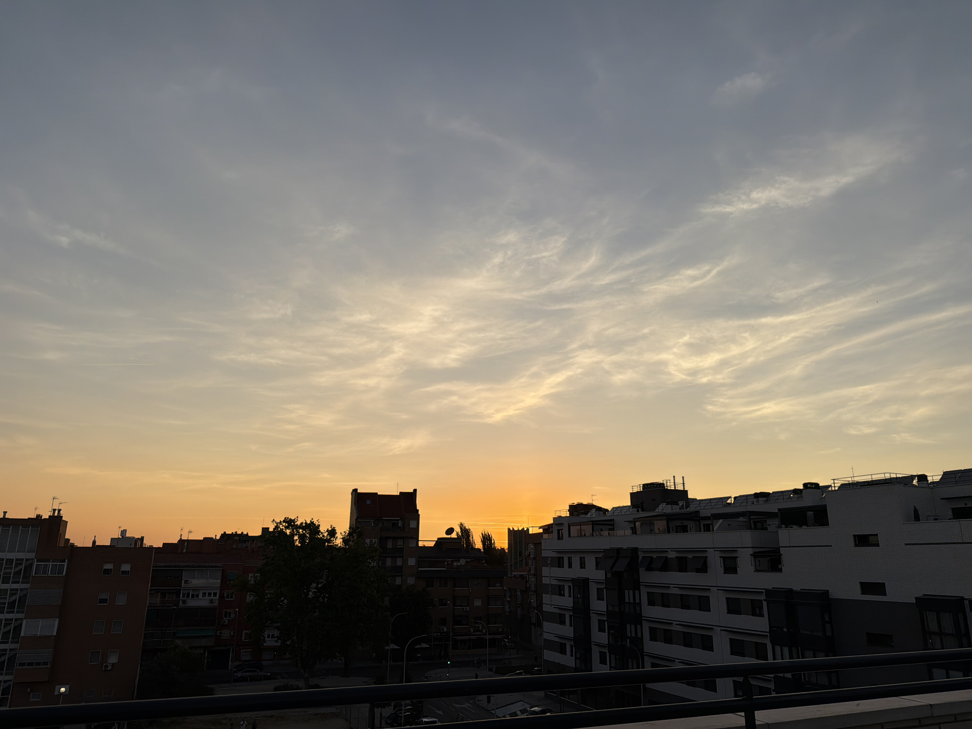 The image captures a cityscape at dusk, with the sun setting on the horizon, casting a warm orange glow across the sky. Wispy clouds are scattered across the sky, some tinged with the colors of the sunset. In the foreground, silhouetted buildings of varying heights line the horizon. The buildings are modern, with some featuring balconies and windows. A dark railing is visible in the very bottom of the frame, suggesting the photo was taken from a high vantage point, possibly a balcony or rooftop. Trees are visible interspersed between the buildings, adding a touch of greenery to the urban landscape. The overall scene conveys a sense of calm and the transition from day to night in a city.