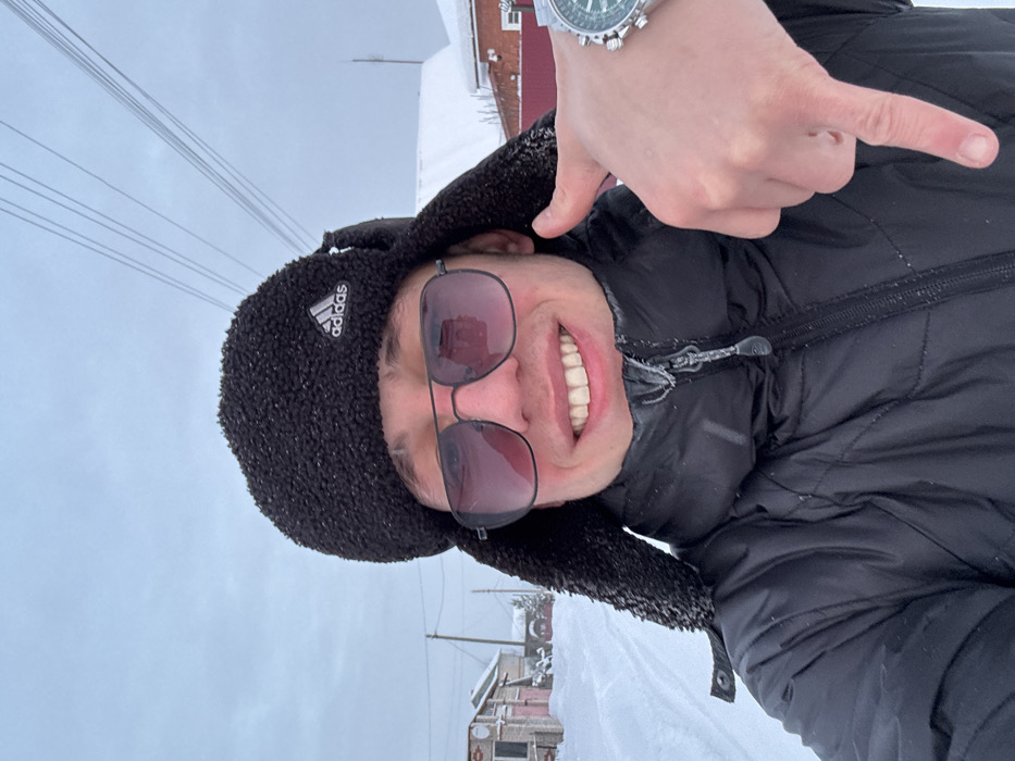 A young male individual, appearing in his late teens or early twenties, is captured in a headshot-style photograph taken outdoors in Ольховка, Russia, during winter. He wears a black Adidas ushanka (trapper hat) and dark-tinted aviator-style sunglasses, reflecting the surroundings. His facial expression is a wide smile, revealing his upper teeth. He is dressed in a black, insulated winter jacket with a visible zippered collar, which shows traces of frost or snow. On his left wrist, a silver-toned wristwatch with a green face is prominently displayed. His right hand is visible in the lower right quadrant, with the index finger extended downwards, pointing towards the bottom of the frame. The background shows a snow-covered environment, characteristic of a street or path in a residential area. Several single-story buildings with snow-covered roofs are visible on both sides of the path, including what appears to be a house with a garage on the left. Utility poles and power lines extend across the upper background against an overcast, light grey sky. The ground is entirely covered in a thick layer of white snow.