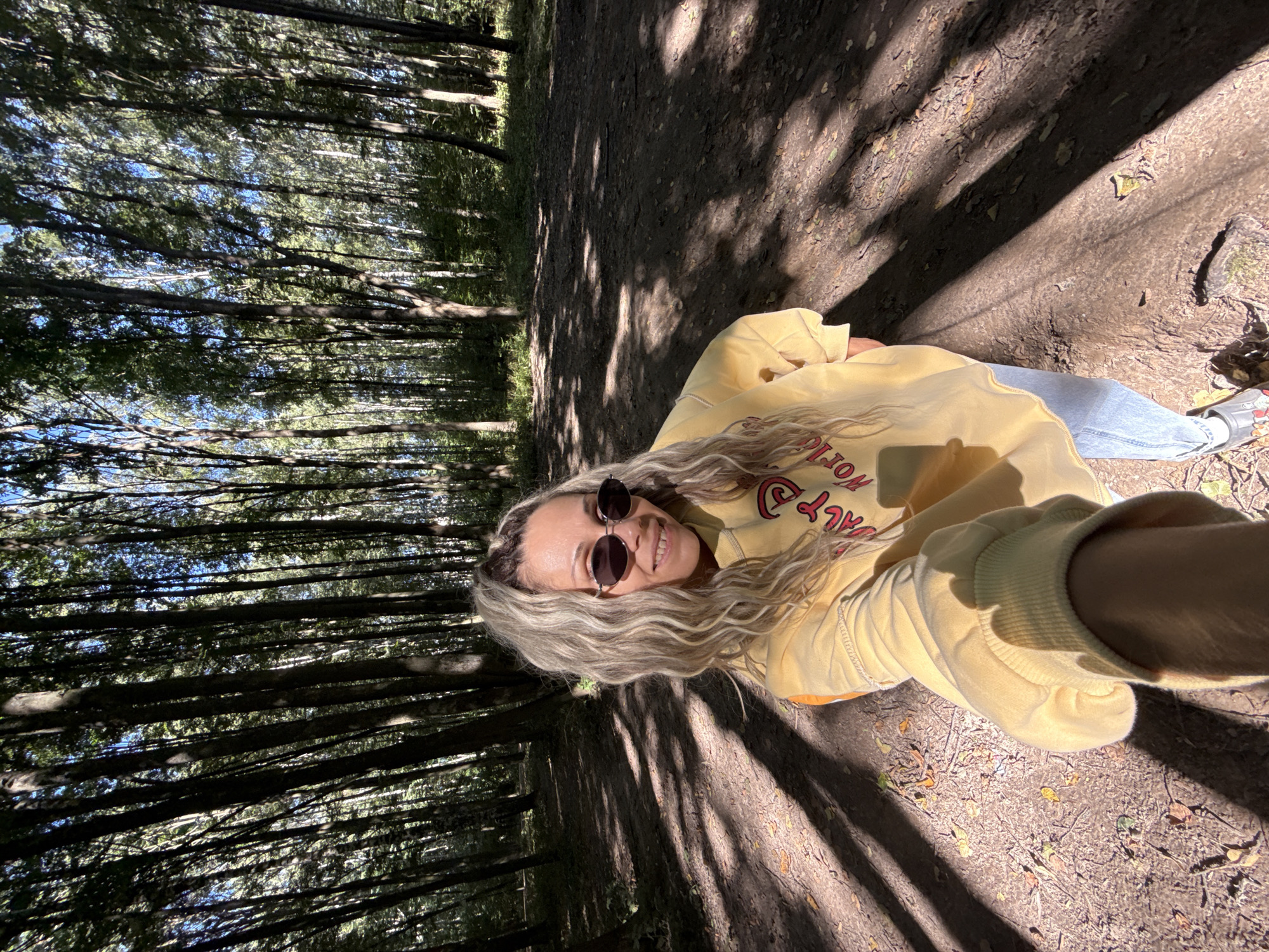 A woman with blonde, wavy hair and sunglasses is taking a selfie in a forest. She is wearing a yellow sweatshirt and blue jeans. The forest is dense with tall trees and sunlight is filtering through the leaves, creating shadows on the ground. The path the woman is on appears to be dirt and covered with leaves. The woman is smiling and looking at the camera.