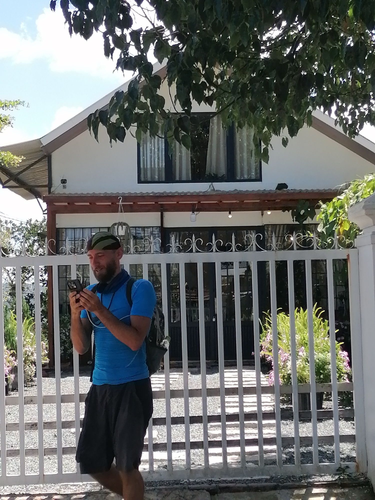 A man with a beard wearing a blue t-shirt, dark shorts, a green and black bandana, and a backpack stands outside a building in Da Lat, Vietnam. He is looking down at his phone, holding it with both hands. In the background, the building has white walls, a brown-trimmed roof, and large windows covered by sheer curtains. A white metal fence with decorative elements is in the foreground, separating the man from the building. Plants and trees are visible around the building and behind the fence. The sky is blue with some clouds.