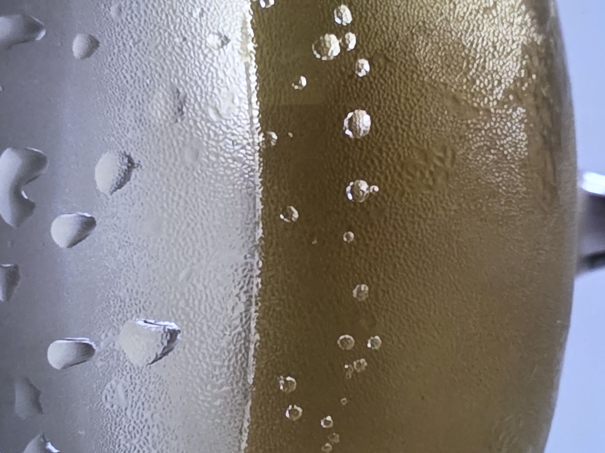 A close-up, macro shot focuses on the condensation and bubbles inside a glass of a golden-hued beverage, likely champagne or a similar sparkling wine. The glass has a textured surface with numerous small bumps, creating a rough, frosted appearance. Water droplets, ranging from tiny specks to larger, teardrop shapes, are visible on the exterior surface of the glass, particularly in the upper portion. The liquid inside the glass is a rich, translucent gold, and it is actively producing small, effervescent bubbles that rise towards the surface. These bubbles are of varying sizes, with some appearing as single spheres and others forming small clusters. The lighting highlights the sheen of the liquid and the clarity of the bubbles, creating a sense of refreshing effervescence. The background is a soft, blurred light blue.