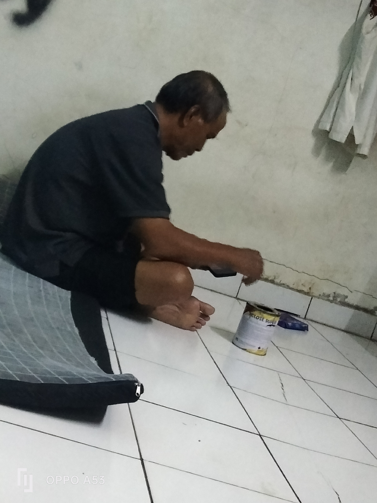 A man is sitting on a tiled floor, hunched over a can of what appears to be paint. He is wearing a dark t-shirt and dark shorts. To his left is a folded sleeping mat with a zipper visible. The wall behind him is light-colored and appears to be cracking in places. A piece of white clothing hangs on the wall to his right. The scene is indoors, in a dimly lit room. The floor is white tile with dark grout lines. The overall impression is one of a simple living space, possibly a room in a house or a dormitory. The presence of the sleeping mat suggests that this might be where the man sleeps. The can of paint and his engagement with it could indicate he is either doing some maintenance work or engaging in a hobby. The image does not provide any specific clues to pinpoint the exact location in Denpasar, Indonesia, beyond it being an indoor setting.