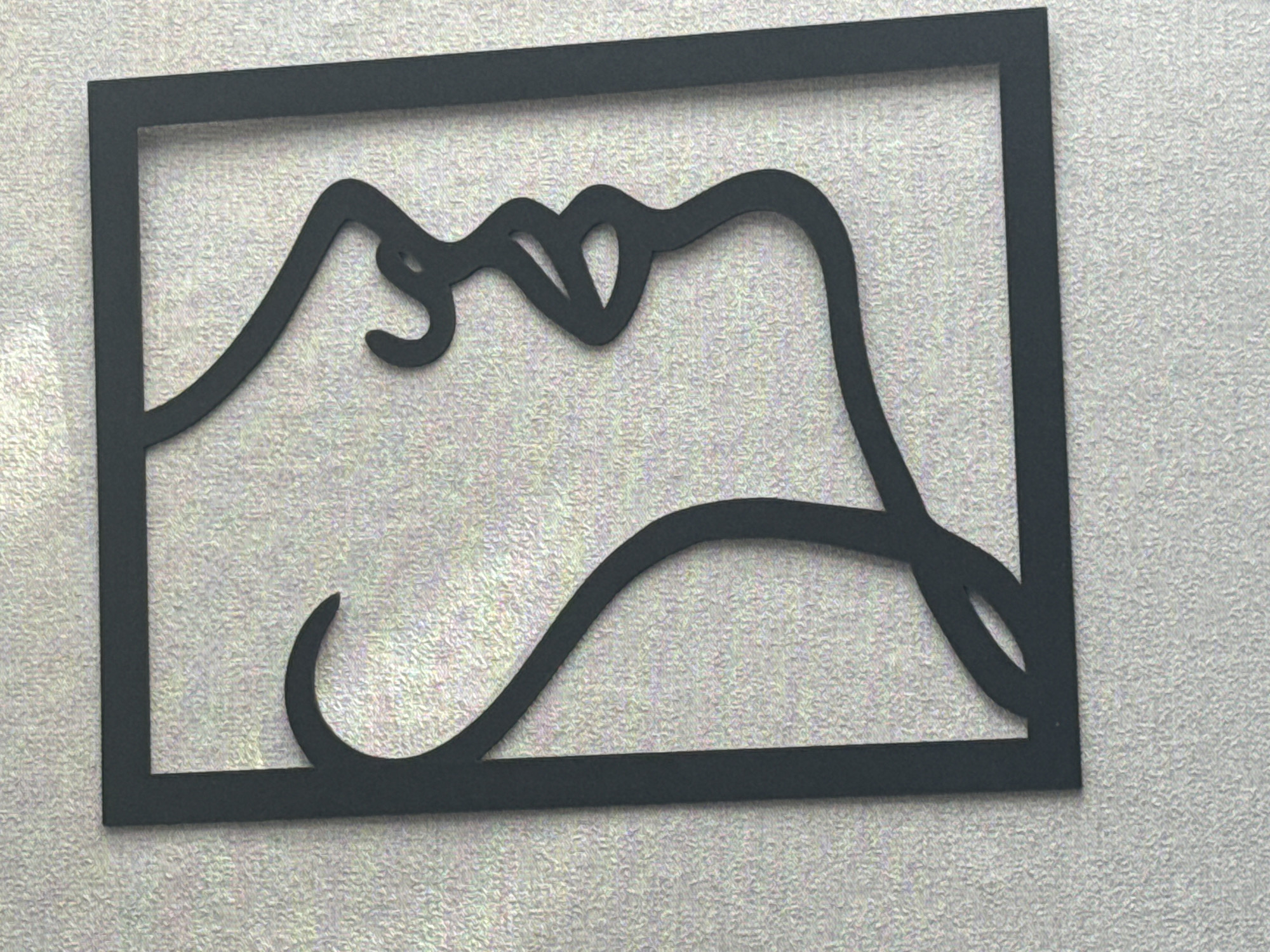 The image displays a black, abstract metal wall art cutout of a woman's profile, enclosed within a rectangular frame. The art is mounted on a textured light gray wall. The profile faces to the right, with a curved line representing the back of the head and neck, flowing into the facial features: a distinct nose, lips, and chin. The artwork is positioned vertically. There is no indication of its location in Taysara, Russia, or any specific city.