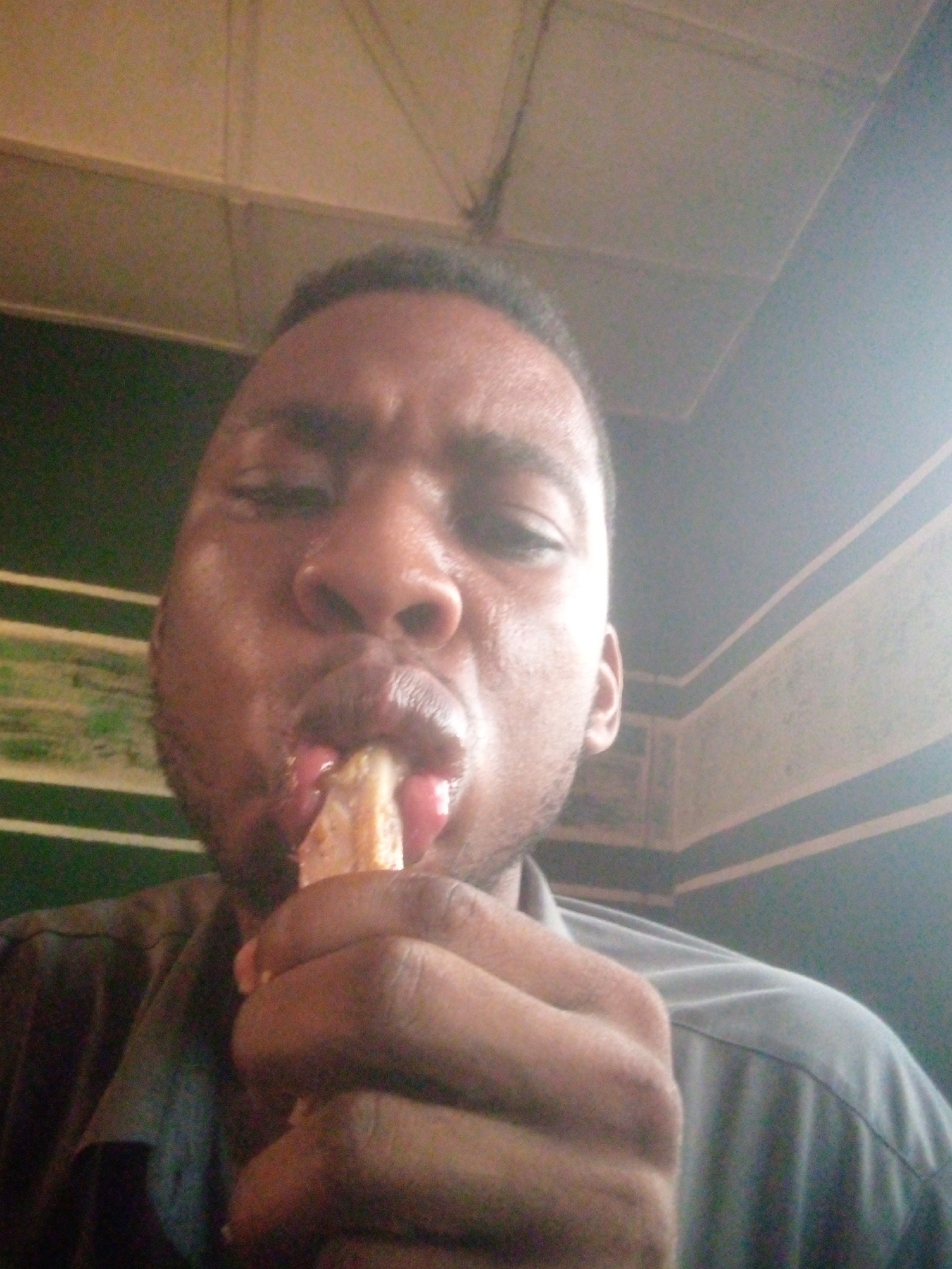 A close-up shot depicts a dark-skinned man eating. He holds a yellowish food item partially within his mouth, with the tips of his fingers visible. The background shows a ceiling and a wall with horizontal stripes. The man is wearing a shirt. Location: Unknown, Unknown.