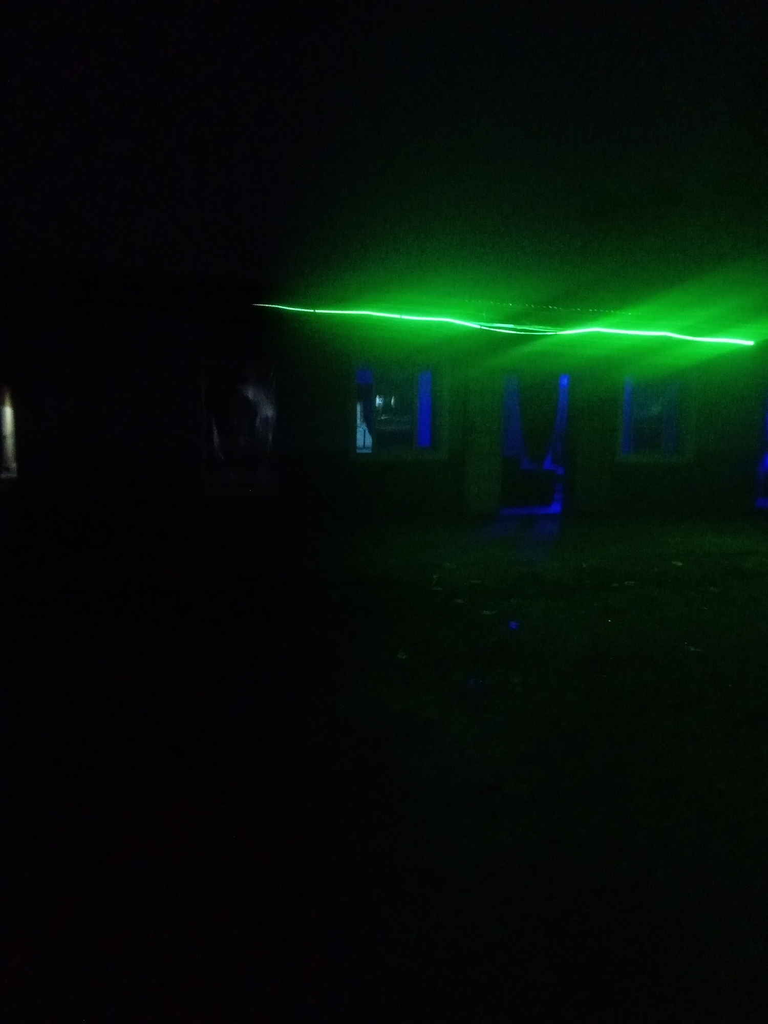 The image depicts a dimly lit, dark interior space, likely an abandoned building or an area with limited illumination, possibly in Sabon Gari, Nassarawa, Nigeria. A prominent, bright green laser beam cuts horizontally across the upper portion of the frame, appearing to emanate from the left and extending towards the right. The laser beam is diffused and slightly blurry, suggesting it is interacting with the air or dust particles in the environment.
In the background, several vertical structures or pillars are visible, illuminated by a blue light. These structures are dark and indistinct, blending with the overall darkness of the scene. The floor is dark and appears to be covered with debris or an uneven surface. The overall atmosphere is one of obscurity and mystery, with the bright green laser providing the only significant source of distinct light. There are no visible people or animals in the image.