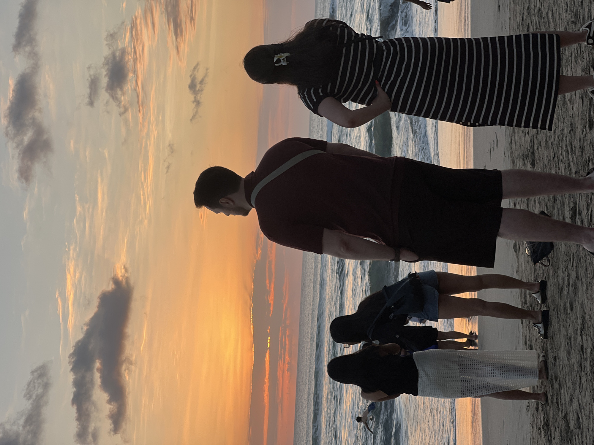 A group of people stand on a sandy beach in Kuta, Indonesia, facing the ocean at sunset. The sky is filled with a gradient of orange and yellow hues, with scattered clouds. The waves are gentle, and the water reflects the warm colors of the sky. In the foreground, a man in a maroon shirt and dark shorts stands with his back to the camera, a strap from a bag slung across his shoulder. To his right, a woman in a black and white striped dress also faces the ocean, her hand on her hip. To the left of the man, two women stand side-by-side. One wears a white, sheer skirt and a black top, and the other wears denim shorts and a black backpack. Further out in the water, a person is swimming. The overall scene is tranquil, capturing a moment of appreciation for the natural beauty of the sunset.