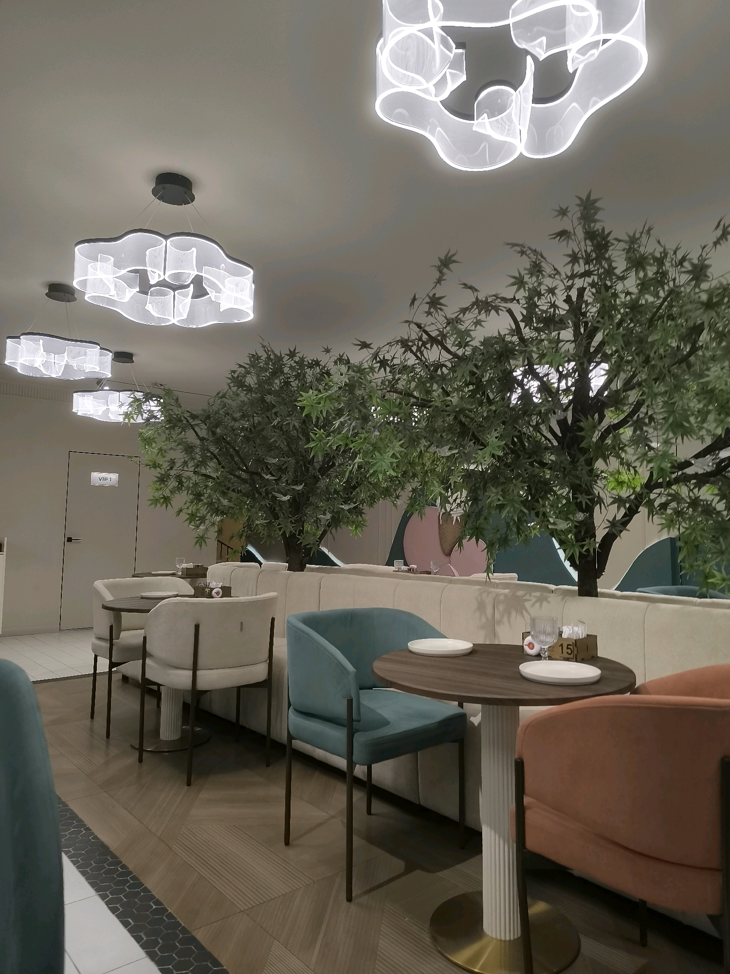 The image depicts the interior of a modern restaurant, likely located in Jezqazğan, Kazakhstan, given the request. The scene is set in the daytime, with artificial lighting from several large, cloud-like chandeliers casting a soft glow. The restaurant features a contemporary design with a light-colored, curved seating banquette. Several tables are visible, some set with plates and glasses. Two faux trees with green foliage are strategically placed within the seating area, adding a touch of nature to the indoor environment. The flooring is a patterned wood-grain tile. A door labeled "VIP 1" is visible on the left side of the image, indicating a private or exclusive area. The chairs surrounding the tables are upholstered in a variety of colors, including a soft beige, a vibrant teal, and a warm orange. No people are present in the image. The overall atmosphere is clean, chic, and inviting.