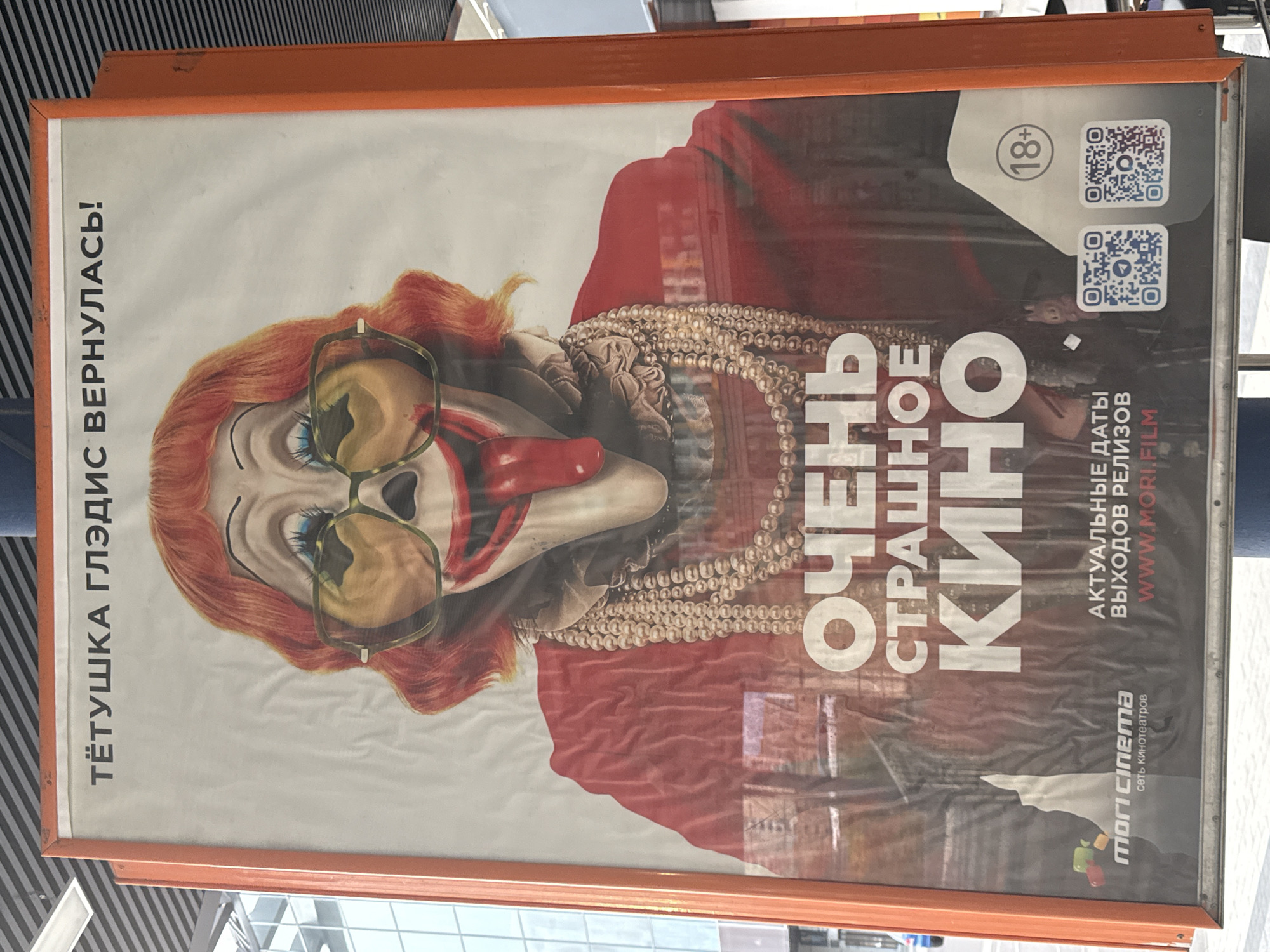 A close-up, eye-level outdoor advertisement poster is framed by an orange metal border. The poster displays a stylized, unsettling image of a clown-like figure with red hair, exaggerated makeup, and large yellow sunglasses. The clown's mouth is wide open, revealing a long, red tongue. The text "ТЁТУШКА ГЛЭДИС ВЕРНУЛАСЬ!" ("Aunt Gladys Has Returned!") is printed in black, bold Cyrillic letters at the top of the poster. Below the clown, large white Cyrillic letters spell out "ОЧЕНЬ СТРАШНОЕ КИНО" ("Very Scary Movie"). Further down, smaller text reads "АКТУАЛЬНЫЕ ДАТЫ ВЫХОДОВ РЕЛИЗОВ WWW.MORI.FILM". In the bottom left corner, a logo for "mori cinema" (сеть кинотеатров) is visible. To the right of the text, there are two QR codes and an "18+" age rating symbol within a grey circle. The poster is affixed to a light grey metal pole. The background suggests an urban environment, with a glimpse of a modern building with reflective windows on the left. The poster itself appears slightly creased and reflective, indicating it is likely protected by a glass or plastic surface. The lighting suggests it is daytime. The image quality is slightly degraded due to resolution and lighting conditions.