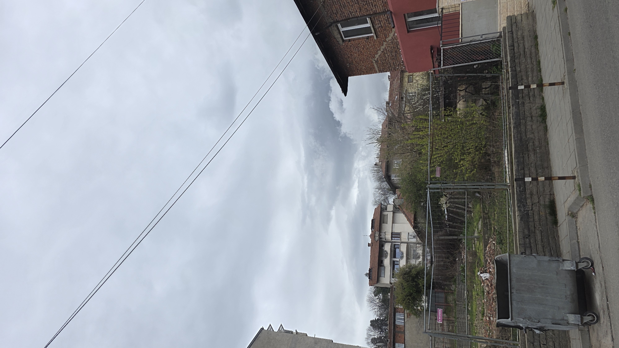 The image depicts an outdoor urban scene, likely in Blagoevgrad, Bulgaria. Overcast sky. Overhead, several electrical wires are visible, stretching diagonally across the frame. Buildings of varying heights and architectural styles line the street. A mix of brick and painted facades can be observed. A metal chain-link fence separates the street from a partially obscured area with greenery, possibly a garden or vacant lot. A metal trash container stands on the edge of the street near the fence. The pavement runs along the right side of the image, with gray concrete slabs. Some foliage (trees and bushes) is seen beyond the fence. The vegetation appears to be in its early stages of growth.