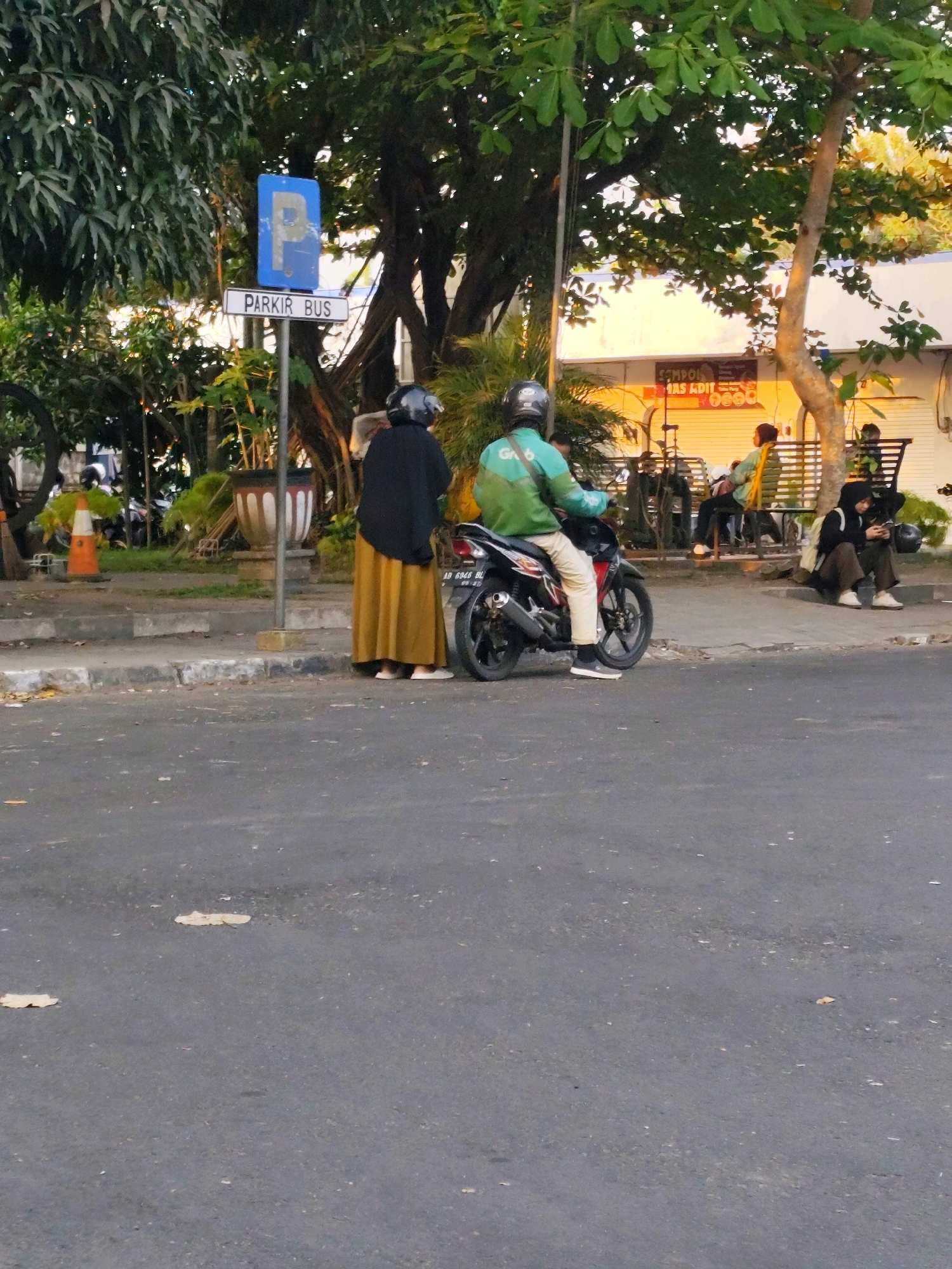 In Bantul, Indonesia, a street scene unfolds under dappled sunlight filtering through trees. A motorcycle rider, clad in a green jacket and beige pants with a helmet, is positioned on a road next to a curb. Beside the rider, a person wearing a black hijab and a long mustard-colored skirt stands, facing away from the camera. A blue parking sign with a white 'P' symbol and the text "PARKIR BUS" is mounted on a metal pole to the left of these individuals. In the background, other people are visible sitting on a bench and on the ground near a building with a bright orange sign that appears to read "SAMPODE AS ADIT." The environment includes asphalt road, a concrete sidewalk, and lush green foliage.