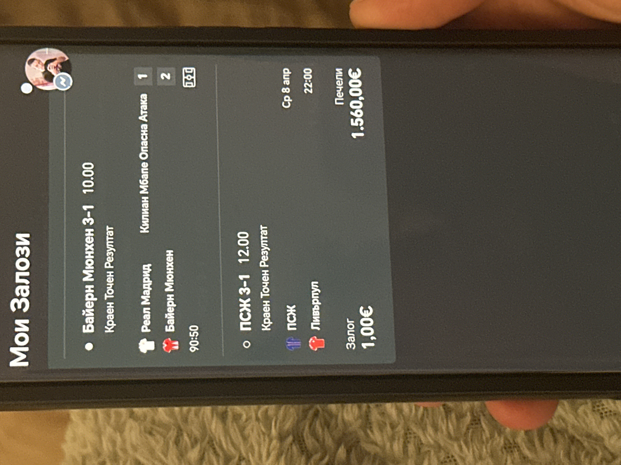 A close-up photograph depicts a cell phone screen displaying a betting slip. The screen shows two bets placed: one for "Bayern Munich 3-1" with odds of 10.00 and a secondary bet for "PSG 3-1" with odds of 12.00. The bet slip indicates a total win of 1,560.00€ on a 1.00€ bet. The date is listed as Wednesday, April 8th, 22:00. The language on the screen is Bulgarian. The location, Sofia, Bulgaria is not evident in the image.