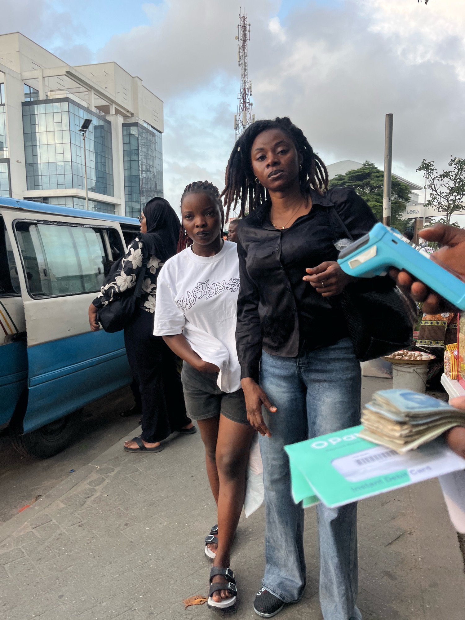 In Lekki Phase I, Nigeria, two women are captured in a street scene next to a blue and white bus. The woman on the left, wearing a white t-shirt and dark shorts, stands beside the open bus door. To her right, a woman with dreadlocks, dressed in a black satin shirt and blue jeans, holds a blue electronic payment device. A hand extends into the frame from the right, offering a stack of cash and a green card. In the background, a modern building with glass facades and a cellular tower are visible under a cloudy sky. The ground is paved with interlocking bricks, and various market stalls are suggested to the right of the frame.