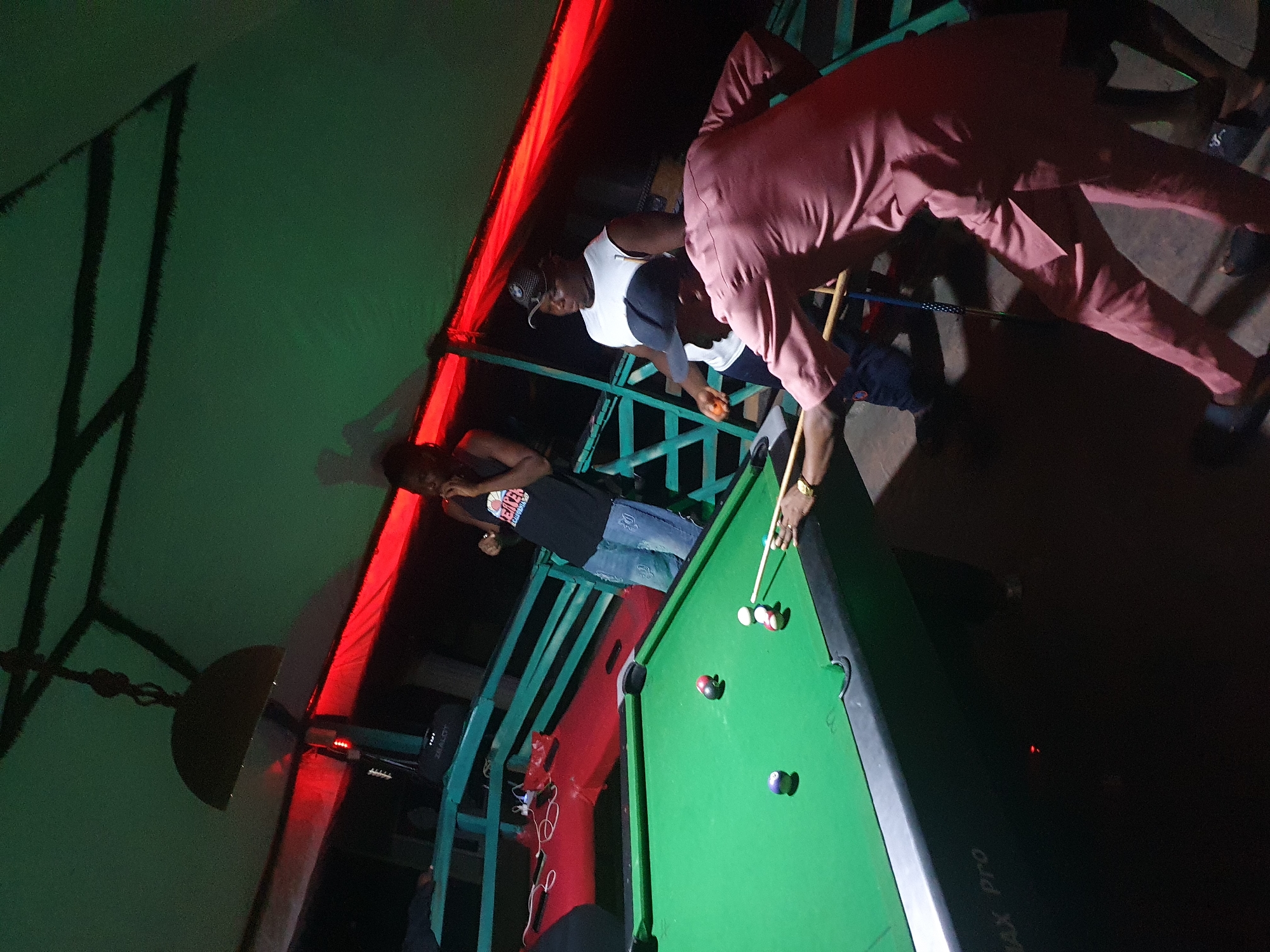 People are playing pool at a venue in Lokoja, Nigeria. There are three people standing on a raised platform near a pool table. One person in a pink suit is taking a shot, and another person in a white tank top is watching. A third person is standing near the edge of the platform. The scene is lit with red and green lights. There are speakers on the platform.