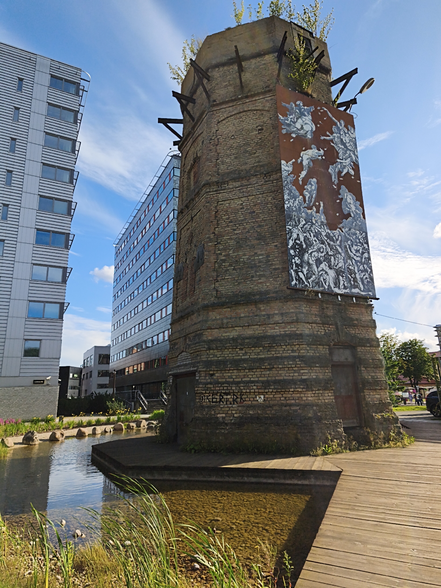 The image captures an outdoor scene with an old brick tower featuring a large mural. Modern buildings are visible in the background. In the foreground, there is a body of water with reeds and a wooden walkway.
The mural on the tower appears to depict a chaotic scene with many figures in monochrome. The tower itself is made of weathered brick and has some green foliage growing from the top. Metal supports are visible extending from the top of the tower.
To the left of the tower, a tall, modern building with a facade of horizontal lines and numerous windows dominates the frame. Further back, another modern building with a glass facade and red accents can be seen.
The water in the foreground has a calm surface, reflecting the sky and surrounding structures. Rocks and reeds line the edge of the water. The wooden walkway is constructed from planks and extends towards the water.
The sky is blue with scattered white clouds, indicating a clear day. The overall impression is a juxtaposition of old and new architecture within an urban environment.
The mural on the tower reads "VÄIKE-TIRGOV" which can be translated from Estonian as "Little Tirgov". This mural is a well-known work of street art and is located on the remains of an old industrial building in the Noblessner Quarter of Tallinn, Estonia.