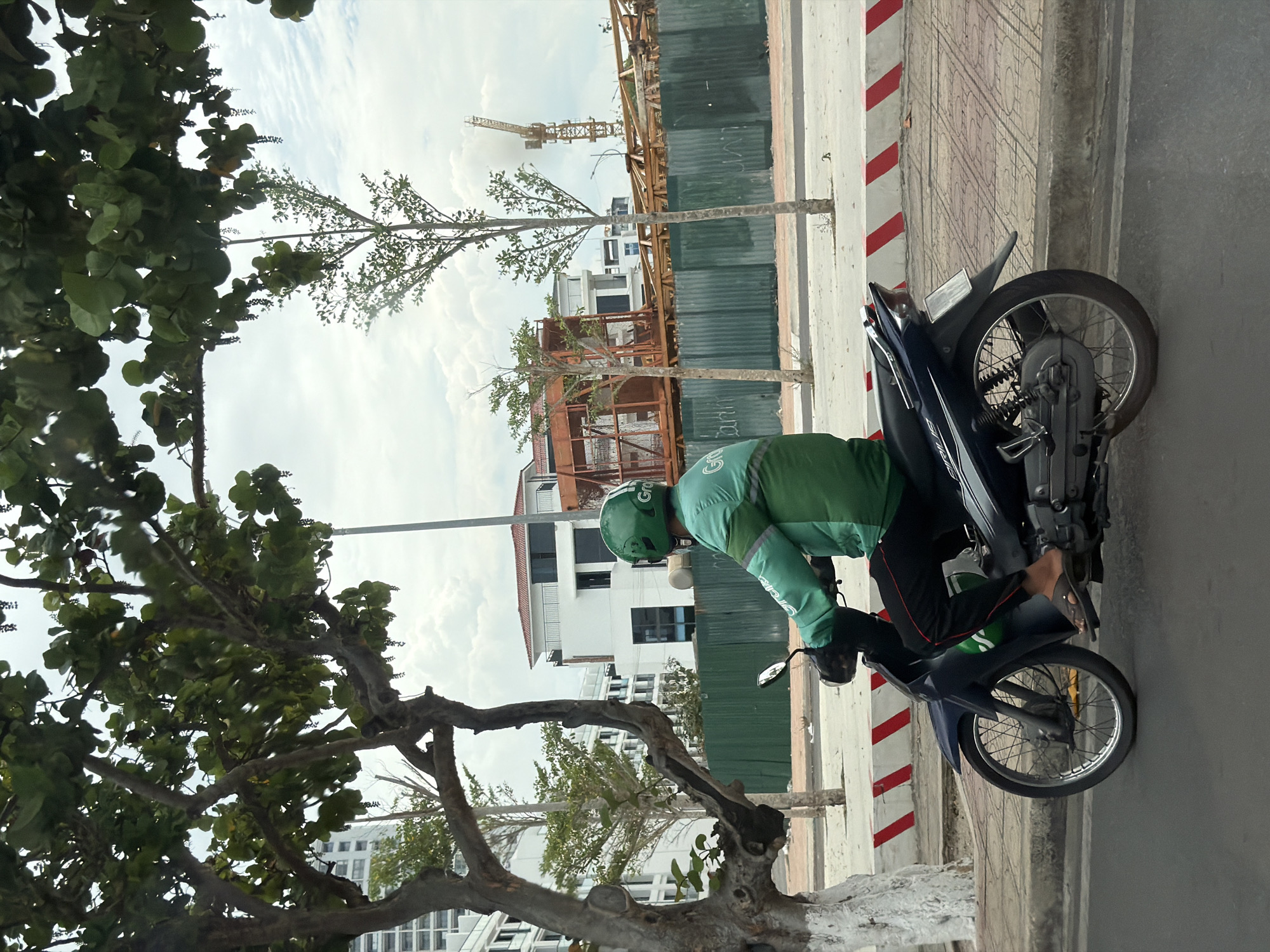 A Grab driver on a blue scooter is parked on a sidewalk in Nha Trang Ward, Vietnam, next to a building construction site with a crane visible in the background. The sidewalk has red and white stripes. Trees with lush green foliage are in the foreground, partially obscuring the scene. The driver wears a green helmet and jacket.