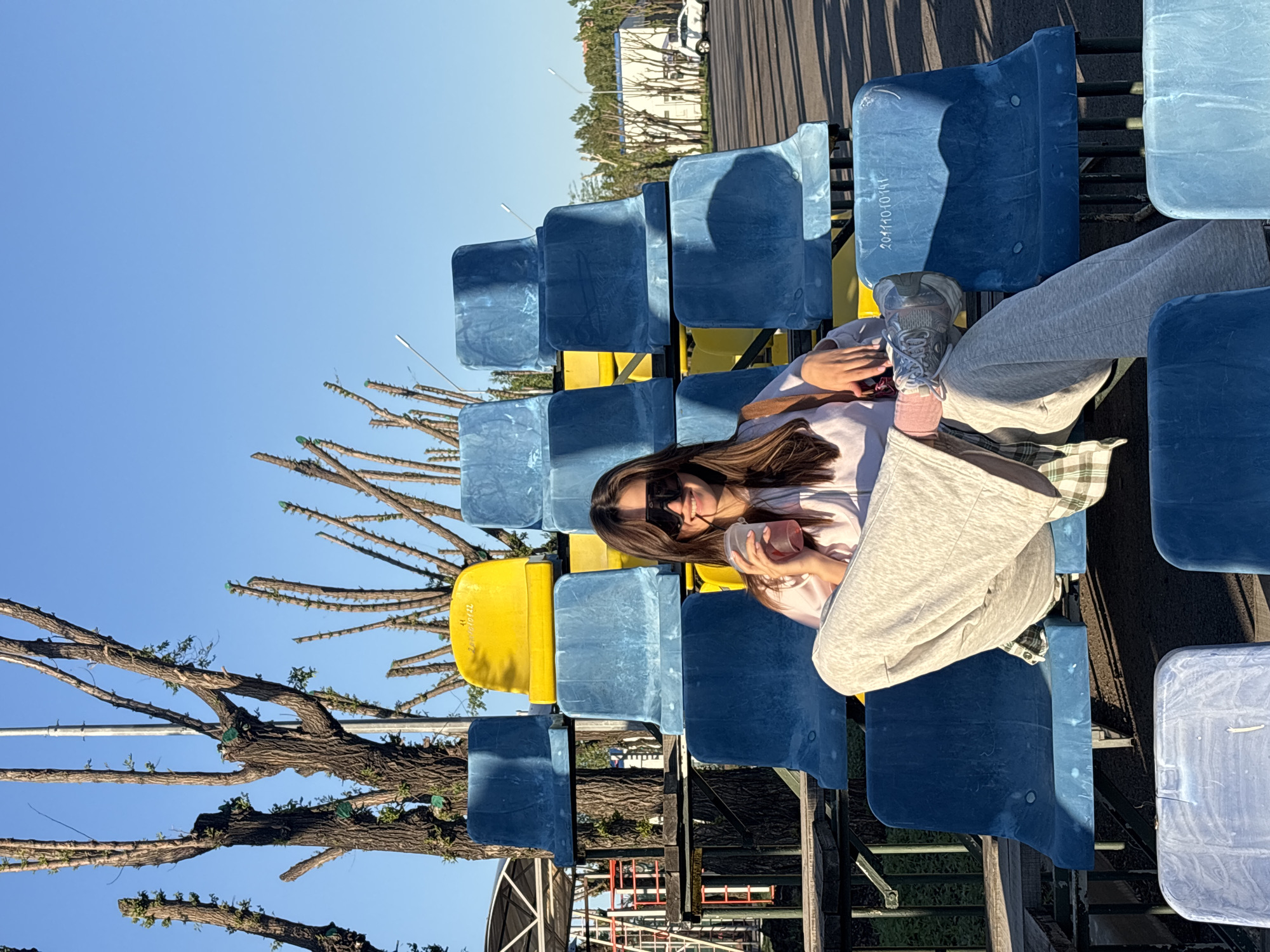 A young woman wearing sunglasses, a white t-shirt, and grey sweatpants is sitting on the blue and yellow seats of an outdoor grandstand. She is holding a cup. The grandstand appears to be near a tree with many branches. In the background are buildings, a street, and a vehicle. The location is Krasnoyarsk, Russia.
