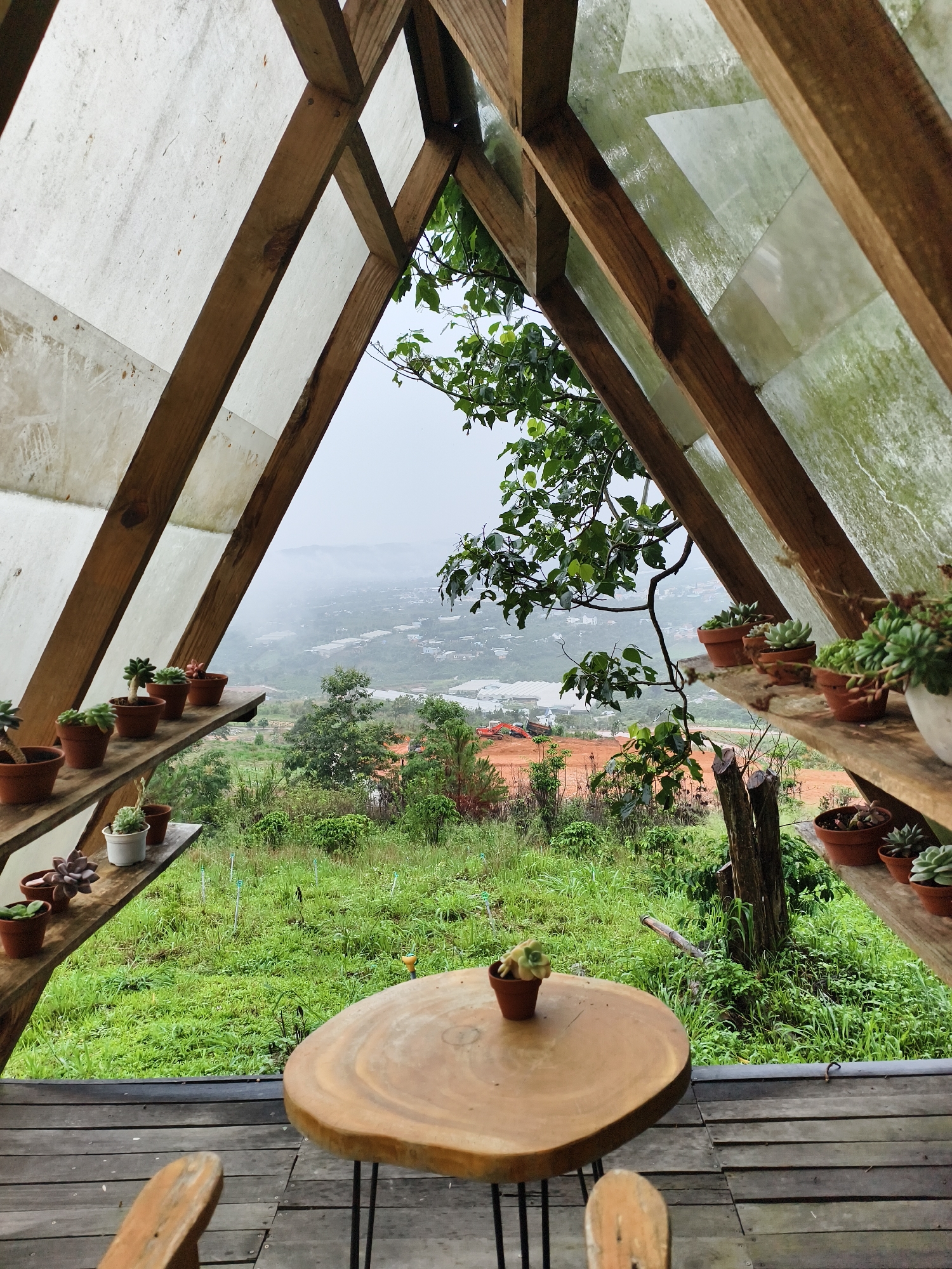 The image depicts a rustic, outdoor seating area, possibly a cafe or garden retreat, with a triangular wooden structure framing the view. The structure is made of rough-hewn wooden beams with translucent sheeting partially covering the roof. Wooden shelves line the sides of the structure, displaying an array of small potted succulent plants. A round, wooden table with a live edge sits in the center of the scene, with a single small succulent pot on top. In the foreground, a wooden deck forms the floor, and the back of a wooden chair is visible.
The view through the triangular opening reveals a lush, green landscape with trees, grass, and a valley in the distance. Buildings and structures are visible in the valley, indicating a town or city. The sky is overcast, suggesting a misty or rainy day.
The location is Unknown, Unknown.