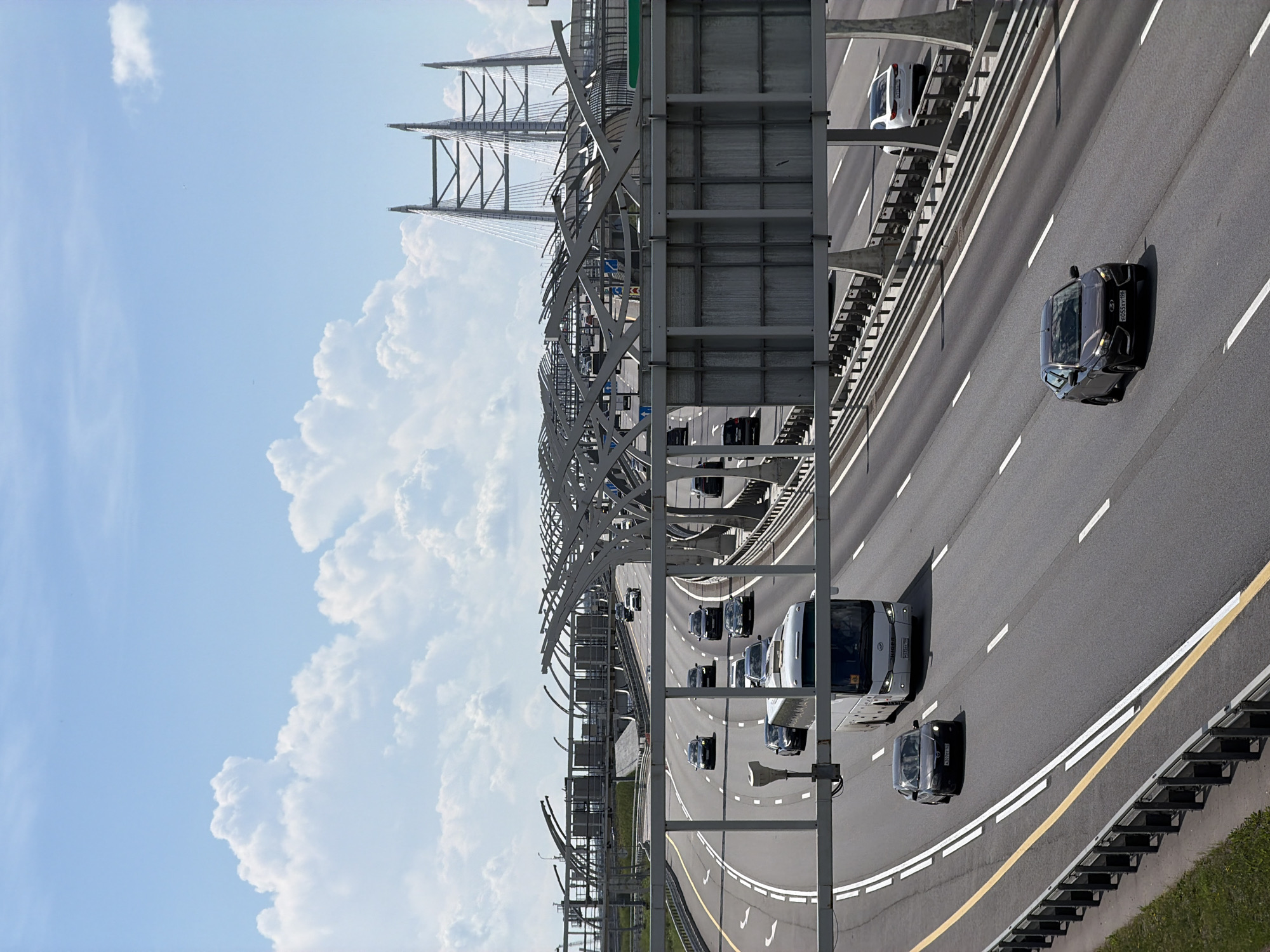 The image captures a view of a multi-lane highway on a bright, sunny day with scattered clouds. A white bus and several cars are traveling on the highway. The highway is flanked by guardrails and overhead structures, some of which appear to be part of a bridge or overpass. In the background, the distinctive pylons of the cable-stayed bridge, likely the Western High-Speed Diameter (WHSD) in Saint Petersburg, Russia, are visible. The overall scene depicts a busy urban transportation artery under a clear sky.