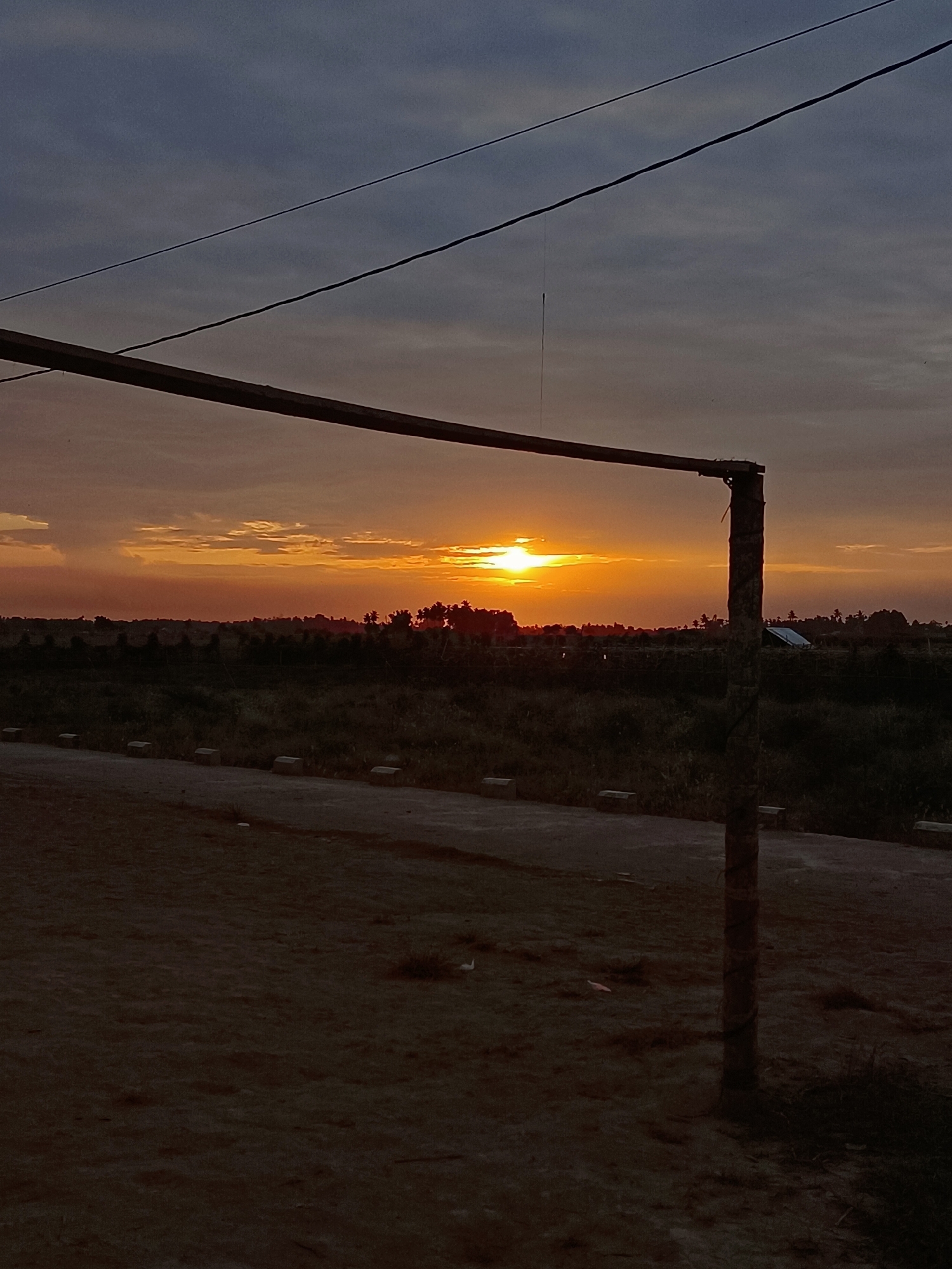 The image shows a sunset over a rural landscape. The sun is setting on the horizon, casting an orange glow across the sky. There are trees and buildings in the distance. In the foreground, there is a dirt road and a fence. The image appears to be taken from a low angle, looking up at the sky. The overall mood is peaceful and serene.
There is no way to determine the specific location in Meunasah Bie, Indonesia from the image alone. However, based on the landscape, it is likely a rural or semi-rural area. The presence of trees and vegetation suggests that it is not a densely populated urban area. The sunset colors and atmospheric conditions could be indicative of the time of year, but without further information, it is impossible to say for sure.