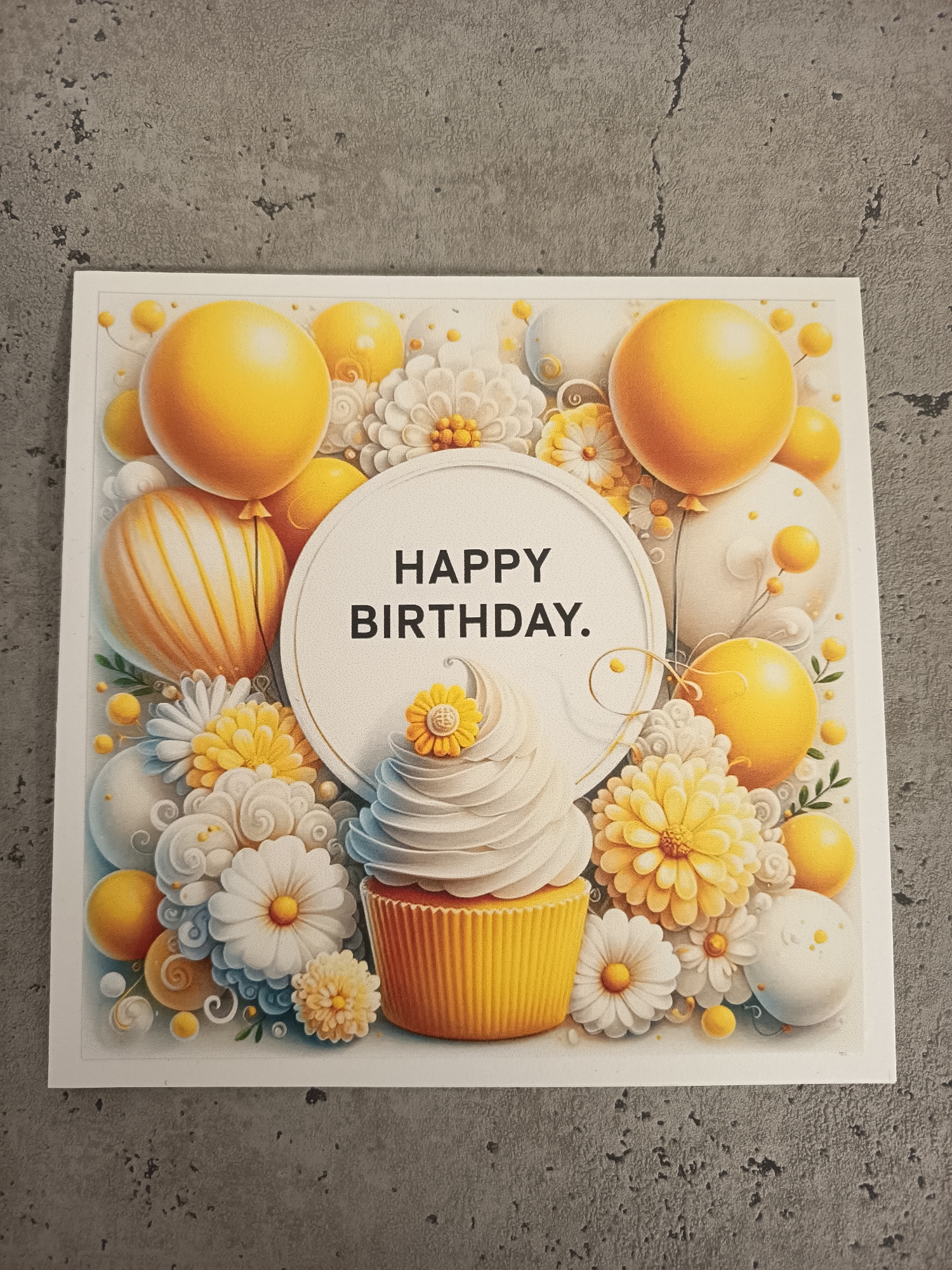 The image is of a square, white "Happy Birthday" card. The card features an illustration with yellow and white balloons, flowers, and a cupcake. "HAPPY BIRTHDAY." is written in black in the center of a white circle. The card is lying on a gray surface. The location is not identifiable as Wangen bei Olten, Switzerland, based on the visible information.