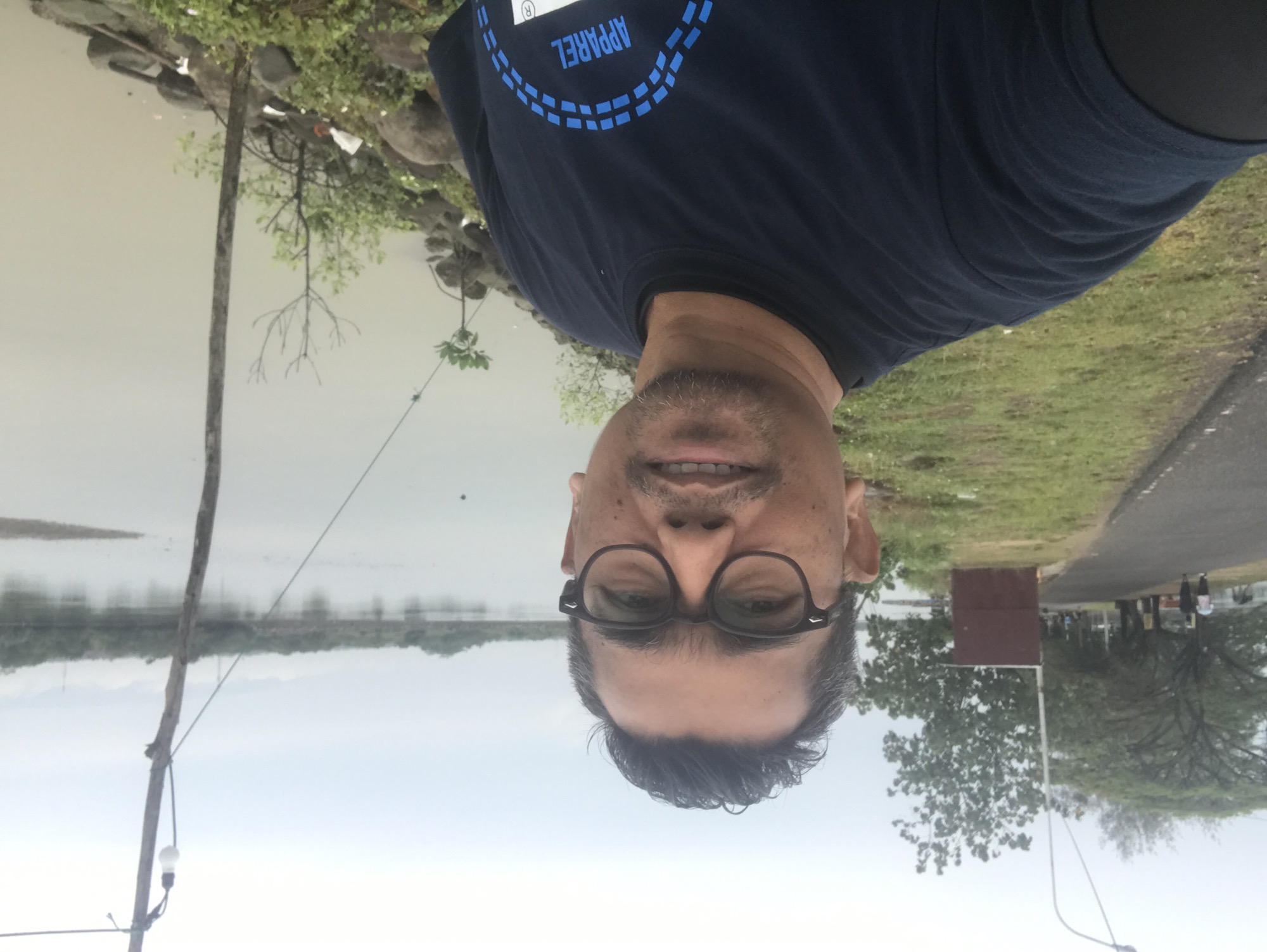 A medium shot photograph depicts a man with glasses and a dark t-shirt standing outside. In the background is a body of water, likely a lake or reservoir, and a tree-lined shore. A road and grassy area are visible on the right side. Based on the location, this is likely taken near the Waduk Kota Lhokseumawe in Indonesia.