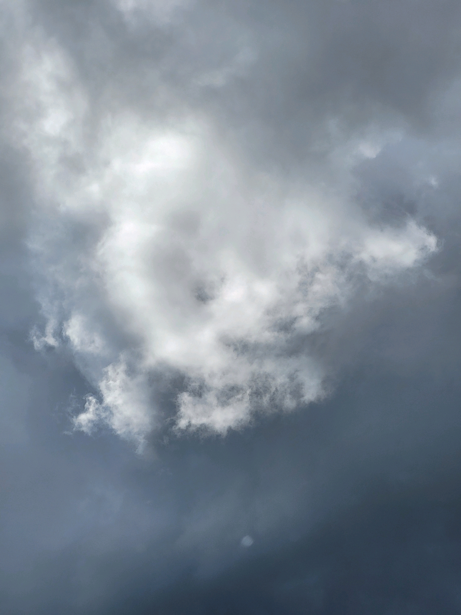The image is a close-up, low-angle shot of a sky filled with dramatic, turbulent clouds. The lighting suggests either dawn or dusk, or possibly a storm brewing, with patches of bright white light breaking through darker, grayer clouds. There are no people, subjects, or objects visible in the frame, only the natural phenomenon of the sky. The image does not contain any specific landmarks or details that would allow for identification of a location in Jelgava, Latvia, or any other specific geographical place. The scene is entirely focused on the atmospheric conditions of the sky.
