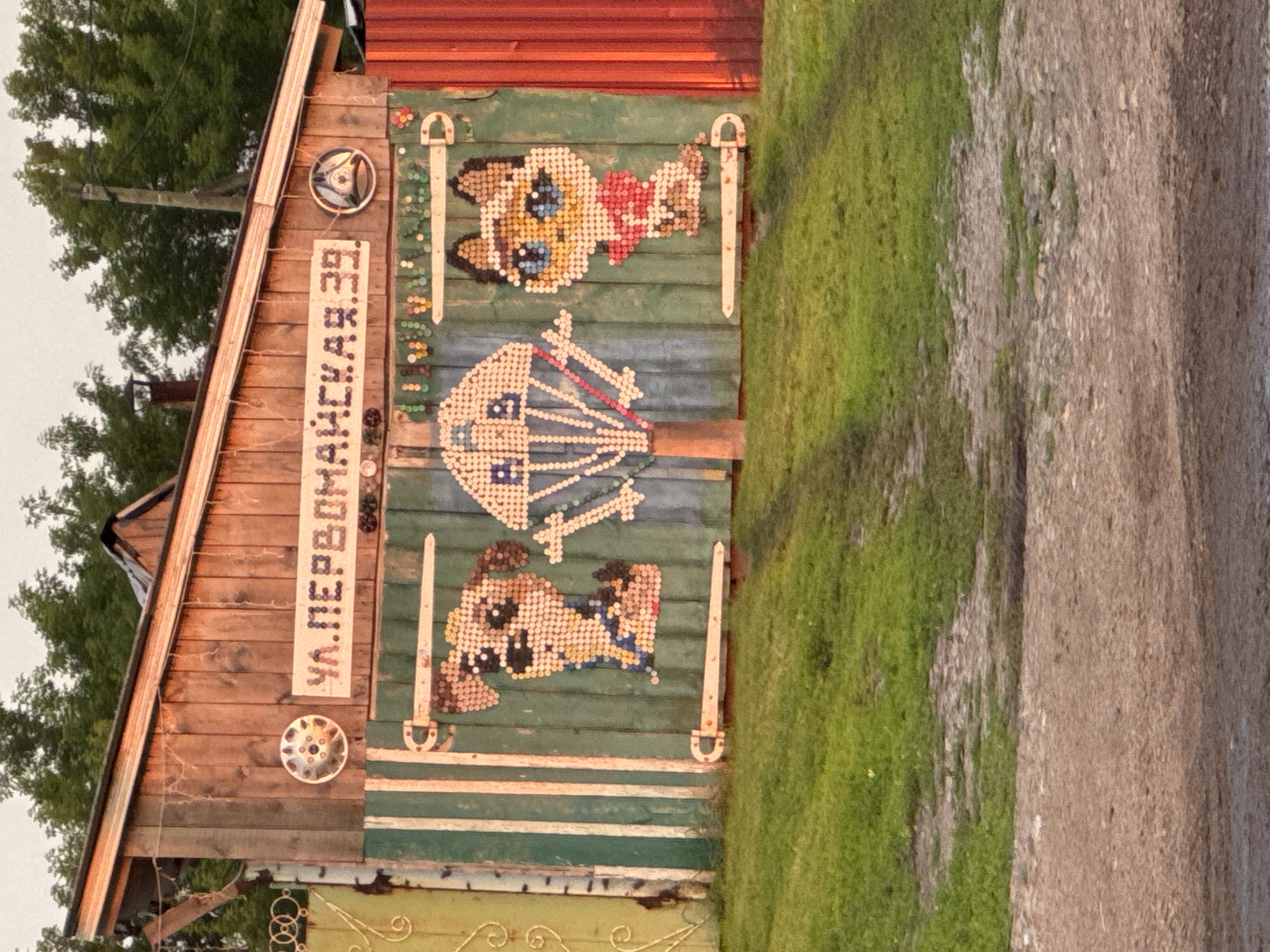 The image depicts a rustic wooden fence adorned with a pixelated mosaic depicting a dog, a parachute, and a cat. Above the mosaic, a sign in Cyrillic reads "УЛ.ПЕРВОМАЙСКАЯ. 39," indicating "Pervomayskaya Street, 39." The fence is situated in the city of Бигила, Russia. The scene is illuminated by natural light, casting shadows on the grassy foreground and a dirt path. The surrounding environment suggests a rural or semi-rural setting, with trees visible in the background. The fence itself appears to be constructed from wooden planks, with metal hinges and decorative elements.