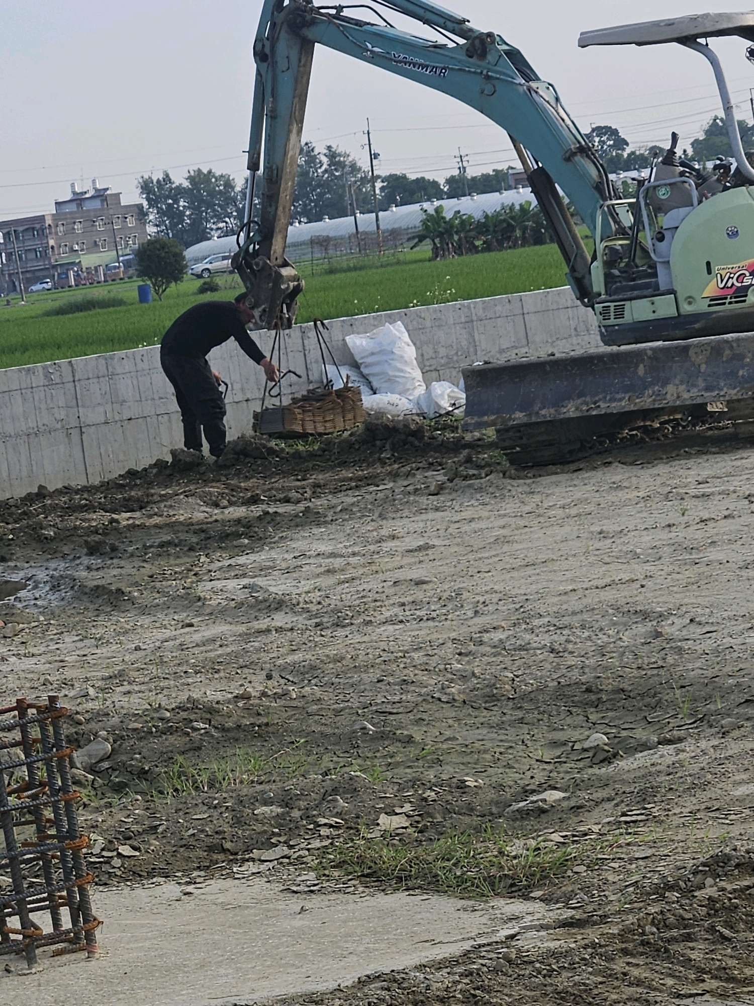 In Xikou Township, Taiwan, a construction scene unfolds. A person, dressed in black, is bent over near a crane, appearing to be handling some sort of netting or material. The crane itself is a teal and green excavator, with the brand name "YANMAR" visible on its arm. To the right of the excavator is a large blade or shovel attachment. In the background, a field of green crops stretches towards a building with multiple windows and a distinctive roofline. Beyond the field, a white car is parked. The ground in the foreground is uneven, composed of dirt, mud, and some scattered rocks. A metal rebar cage sits to the left of the frame. There are also white bags of material, possibly sand or cement, near the concrete barrier. The sky is overcast, suggesting a hazy or cloudy day.
