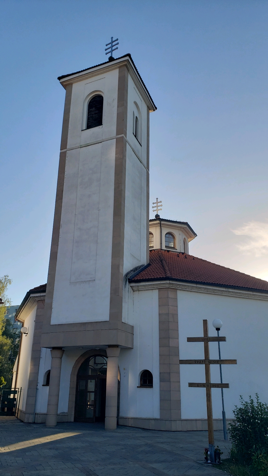 The image shows the exterior of a white church with a tall bell tower, a red-tiled roof, and arched windows. A wooden cross with inscriptions stands in front of the church, to the right of the main entrance. The sky is clear blue with some faint clouds. The church is likely located in the city of Prešov, Slovakia. The scene appears to be a bright, sunny day with shadows cast on the ground, suggesting it was taken in the late afternoon. There are no people visible in the image.