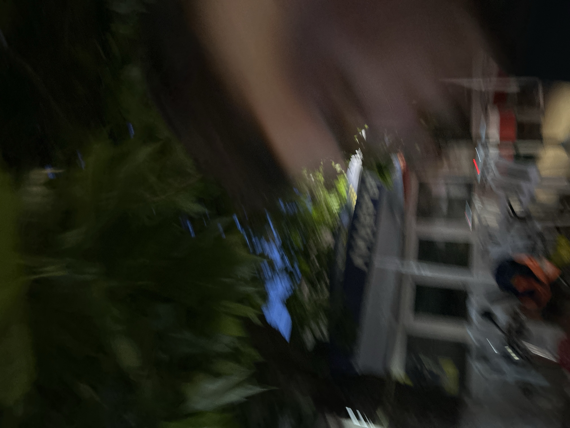 The image presents a highly motion-blurred and low-light outdoor scene, hypothetically situated in Dzhebel, Bulgaria, though no discernible landmarks confirm this specific location. The lighting conditions suggest late dusk or nighttime.
The upper left quadrant of the image contains heavily blurred, dark green foliage, indicating trees or dense vegetation overhead, with fragments of a dark blue sky visible through gaps in the canopy.
In the mid-to-lower left, a light-colored building or structure featuring several indistinct, vertically oriented window-like apertures is discernible. Directly in front of this structure, multiple blurred objects consistent with outdoor seating, such as chairs and tables, are present. Some of these objects exhibit faint red glowing elements. A darker, amorphous shape, potentially a parked vehicle like a scooter or bicycle, is partially visible in the bottom left, accompanied by a blurred orange object.
The entire right side of the frame is occupied by a large, featureless, blurred mass ranging in color from tan to dark brown. This mass, which extends from the top to the bottom of the image, appears to be a close-up foreground obstruction, possibly part of a person or a large object accidentally entering the frame, causing significant obscurity to that portion of the scene. The bottom right corner is profoundly dark, possibly due to a shadow cast by this foreground element.
The pervasive motion blur across the entire image indicates that the camera was in motion during the exposure.