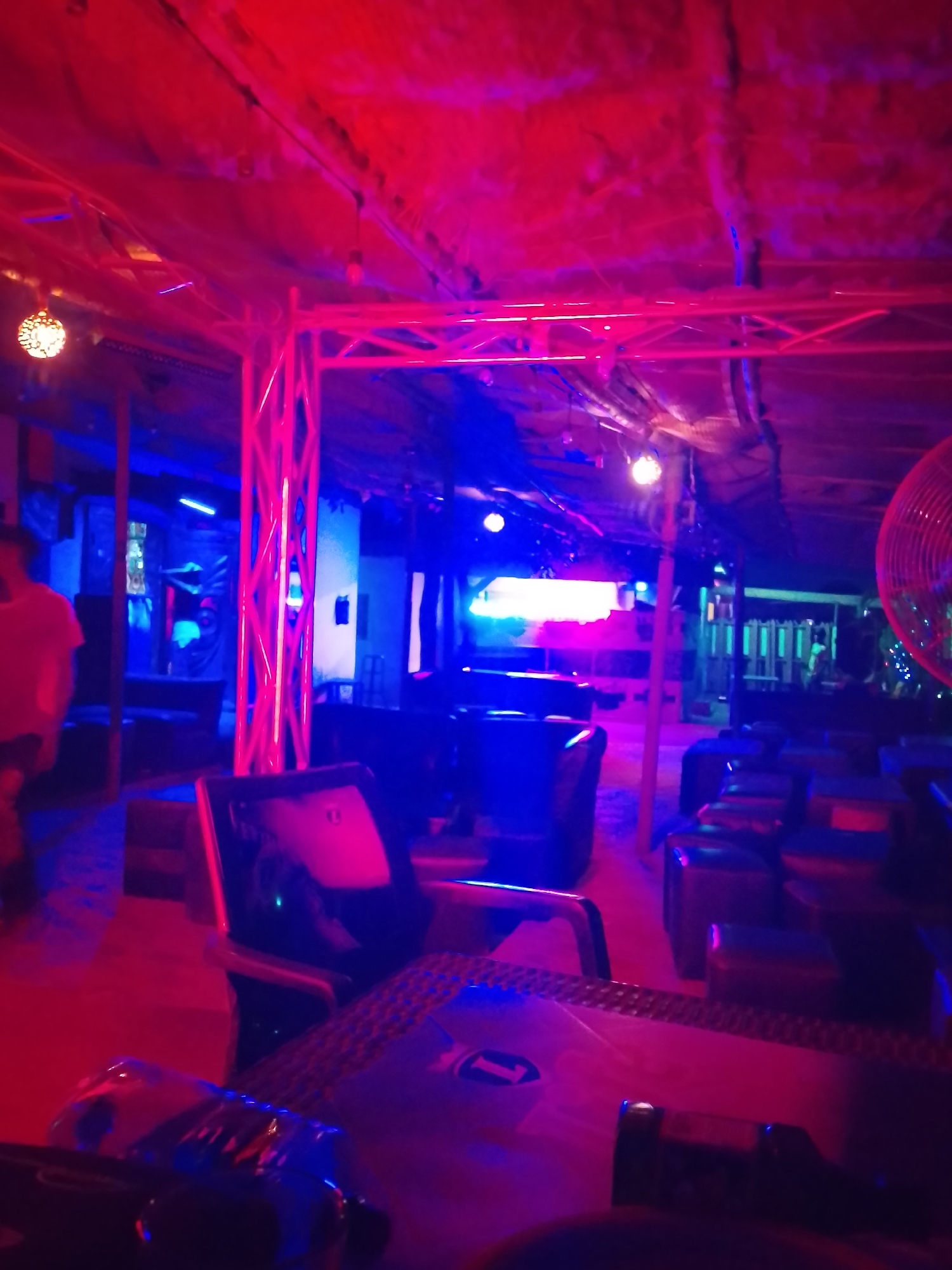 The image depicts an indoor or semi-covered lounge or club environment, characterized by strong, atmospheric red and blue lighting. The overall scene is dim, with artificial lights casting vibrant hues across the space. The ceiling appears to be made of a textured, possibly fabric or thatched, material, bathed in a deep red glow.
Structural elements, such as metal trusses and support beams, are visible throughout, also illuminated in vivid red and blue. Various seating arrangements are present, including dark-colored chairs and low tables or ottomans arranged in clusters, suggesting areas for socializing. In the foreground, a dark table with a textured, possibly metallic, surface is visible, reflecting the red light. On this table, a small circular object with the number "1" clearly marked is present. What appears to be a glass bottle, reflecting blue light, is also on the table.
On the far left, a blurry figure of a person, likely male, is partially visible, wearing a light-colored top that appears reddish due to the ambient lighting. Other indistinct figures might be present in the background. In the mid-ground, a chair with a faint white "H" or similar symbol is discernable. Decorative lighting includes spherical pendant lights and linear blue strip lights. On the right side of the frame, part of a large fan with red blades or housing is visible. The prominent artificial lighting indicates a nighttime setting or an interior designed for an evening ambiance. The scene suggests a relaxed social gathering in a vibrant, modern establishment.