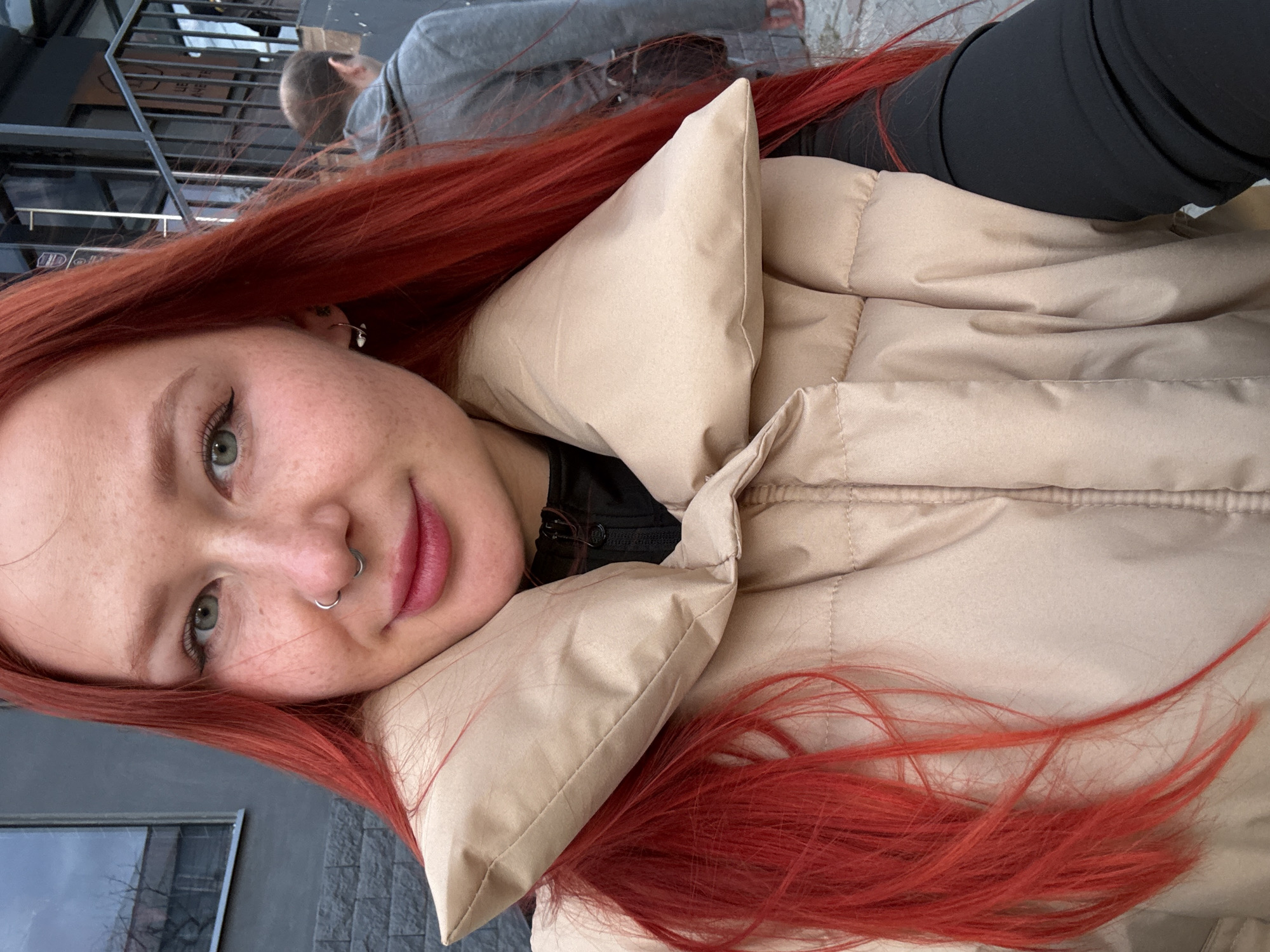 A young woman with striking red hair is captured in a close-up selfie, oriented diagonally in the frame. She has fair skin with visible freckles, green eyes, and is wearing black eyeliner and pink lipstick or gloss. A silver septum piercing adorns her nose, and a small silver hoop earring with a charm is visible in her right ear. She has a subtle, pleasant smile. Her hair is a vibrant reddish-orange, cascading around her face and over her shoulders.
She is wearing a beige, puffy winter jacket and a black top underneath. The material of the jacket appears soft and quilted.
The background suggests an outdoor urban setting, possibly a street or a public area in Rostov-on-Don. Another individual, a male seen from behind, is out of focus in the upper left background, wearing a grey long-sleeved shirt and appearing to walk away. Behind him, there are dark, possibly metallic, vertical bars or fencing, and indistinct building elements. A blurry window reflecting bare tree branches can be seen in the lower left.
The lighting indicates daytime, likely overcast, with soft, diffused light. There is no clearly discernible text in the image; some extremely blurry, unidentifiable characters appear on a sign in the far background of one of the cropped images. The overall impression is a casual, everyday moment.