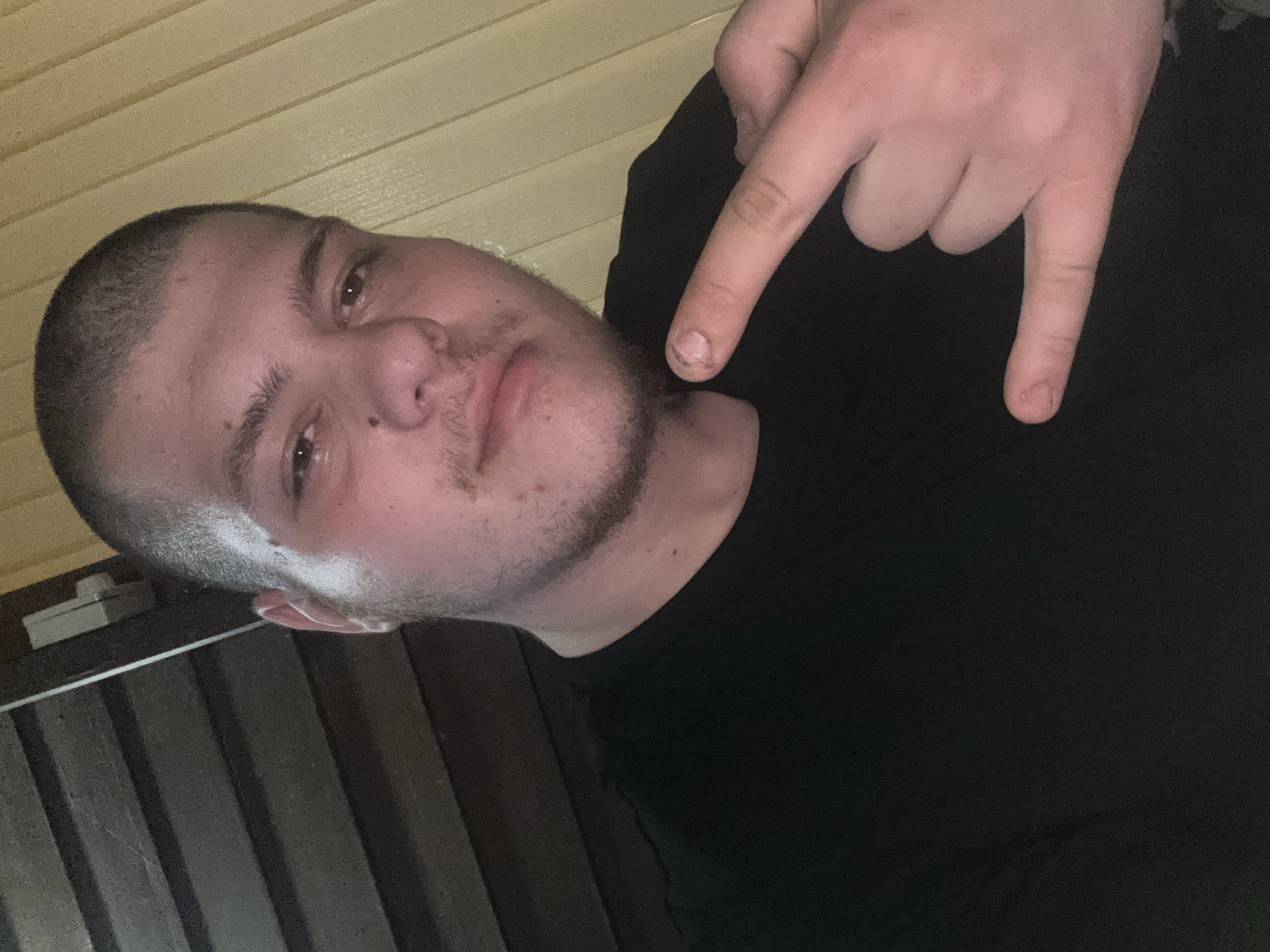A young adult male, appearing to be in his late teens or early twenties, is depicted in a medium shot. He has a very short buzz cut hairstyle and short facial hair growth around his jawline and chin. His face is round, with dark eyebrows and eyes. His skin appears fair but is flushed with red tones, and some minor blemishes are visible. He wears a plain black t-shirt. His left hand is raised in front of his torso, forming a hand gesture commonly known as "rock and roll" or "I love you," with the thumb, index, and pinky fingers extended and the middle and ring fingers curled down. The fingernail on his extended index finger appears short and has visible debris or dirt underneath.
The subject is illuminated from the right, causing the right side of his face and the background behind him to be brighter than the left side. The background is divided vertically. The left side consists of dark brown horizontal slats, possibly wood paneling or blinds, with a small light-colored rectangular object (possibly a switch or fixture) visible at the top left. The right side of the background features vertical light yellow or tan slats or panels. The image appears to be taken indoors or in a covered outdoor area with artificial or diffused light.