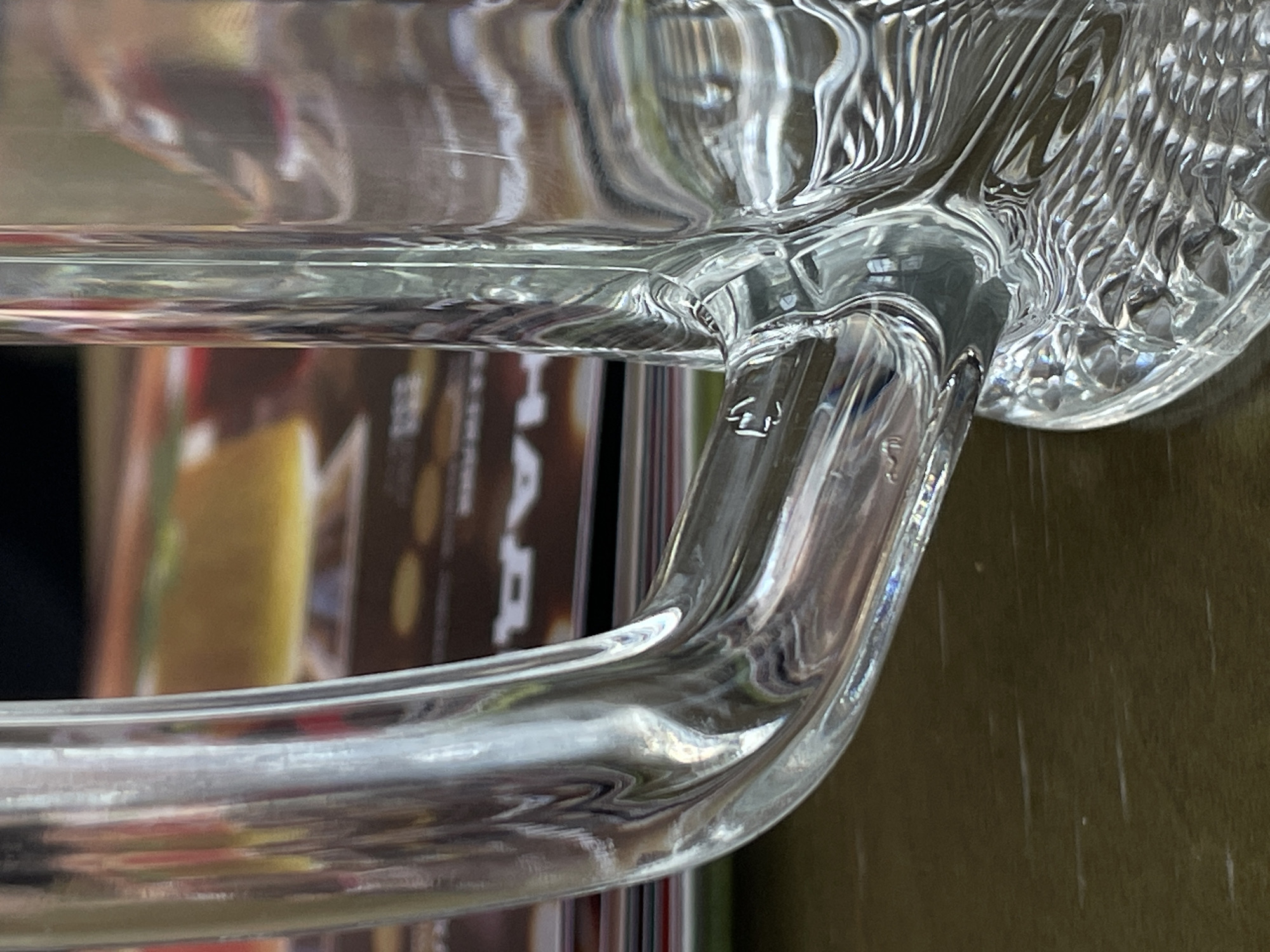 The image shows a close-up, low-angle view of the handle of a clear glass mug. The mug handle is curved and attached to the side of the mug. The glass is reflective, showing distortions of the background which appears to be a stack of books or magazines with colorful covers. The surface of the table beneath the mug is a dark, textured wood grain. There are no people or animals present in the image. The setting is indoors, likely a home or restaurant. The image is a still photograph. The location is not specified in the image itself, and there is no visual information that would allow for its identification as Rostov-on-Don, Russia.