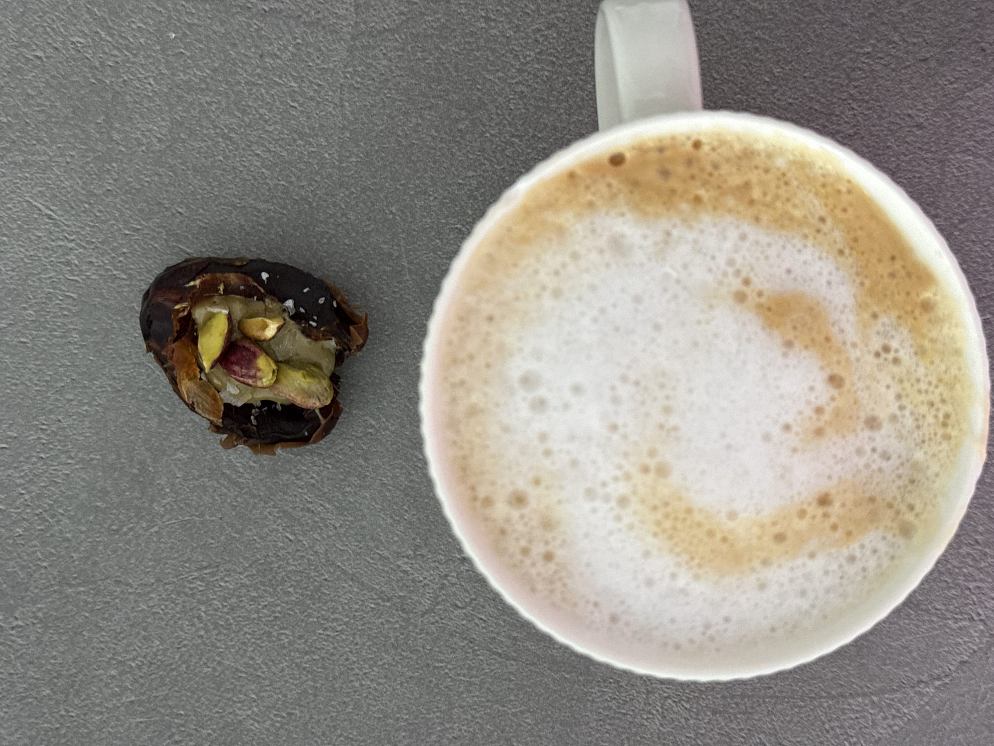 An overhead shot captures a simple arrangement of food and drink on a textured grey surface. To the right, a white ceramic mug, with its handle partially visible, contains a frothy coffee beverage, likely a latte or cappuccino, featuring a thick layer of white foam with light brown coffee art or swirls. To the left, a dark brown, open date is displayed, revealing bright green pistachios inside, some still with their characteristic reddish outer skins. Small white flakes, appearing to be sea salt, are sprinkled over the pistachios and the date. The image depicts a quiet moment, possibly a coffee break with a sweet and salty snack. There are no people or visible text. The scene is located in Ramat Gan, Israel.
