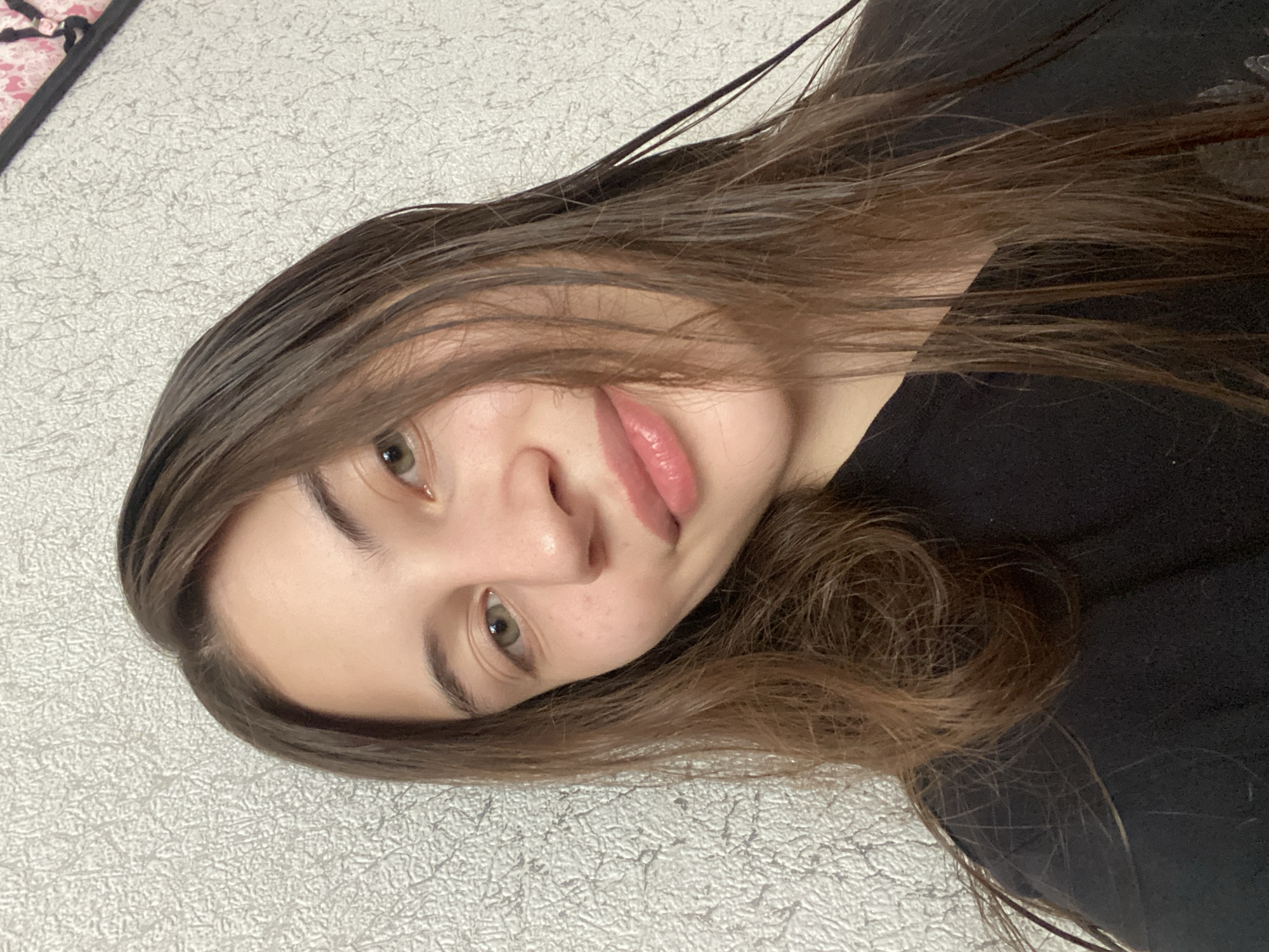 A young woman with long brown hair and light-colored eyes smiles gently at the camera, her head tilted slightly. She is wearing a black t-shirt. The background consists of a textured light-colored wall. The image appears to be a selfie taken indoors, likely in a residential setting. The location is Votkinsk, Russia.