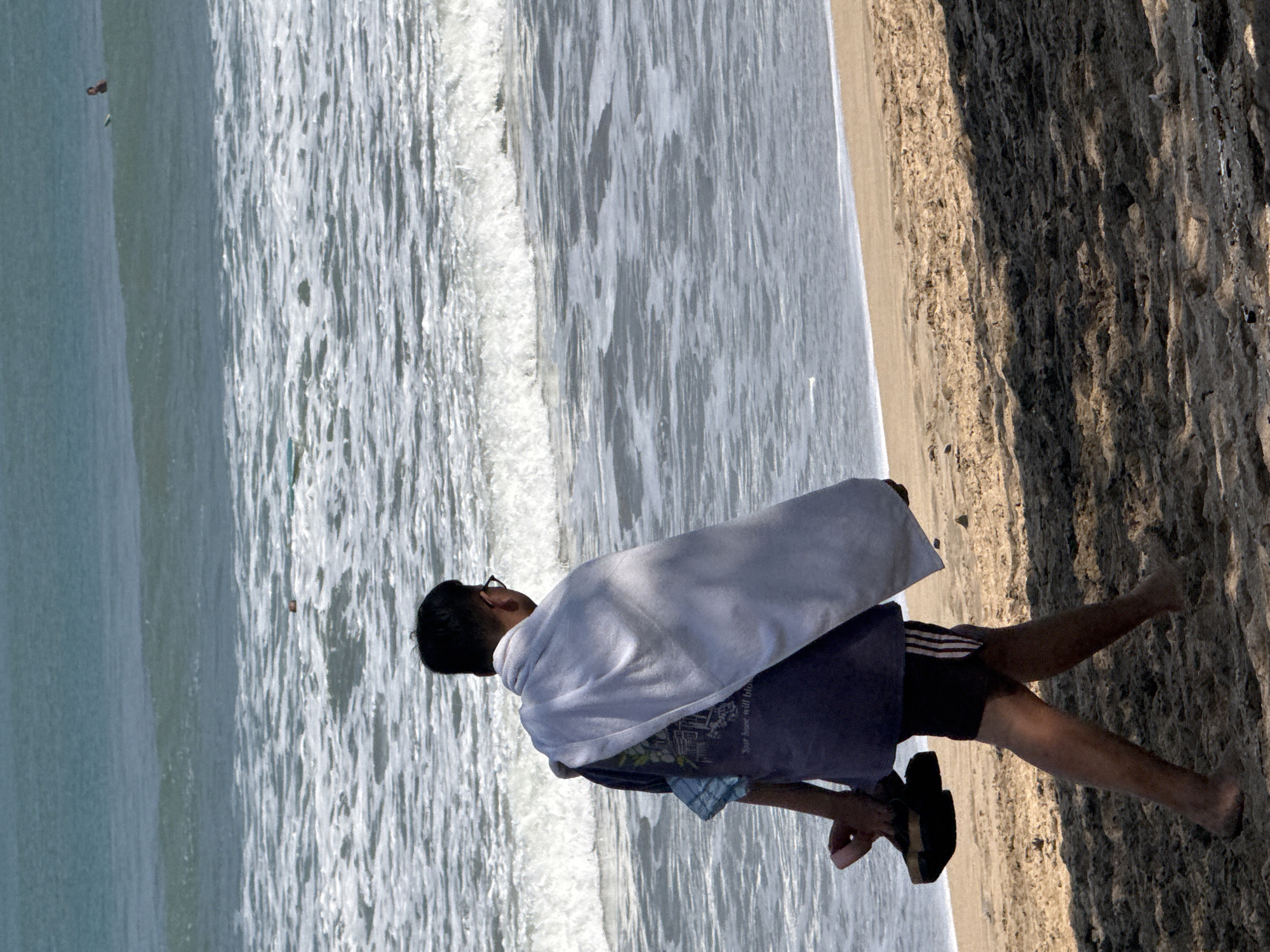A person is walking on the sandy beach in Kuta, Indonesia. The individual, facing away from the camera, is wearing dark shorts with white stripes on the sides, and a dark t-shirt with a graphic print. A light-colored towel is draped over their shoulders and back. They are carrying a pair of dark sandals in their left hand. The beach is wet near the water's edge, with small waves breaking on the shore. In the distance, several people can be seen in the ocean, some with surfboards. The sky is clear and the ocean appears calm. The foreground is a sandy area with shadows cast by an unseen object.