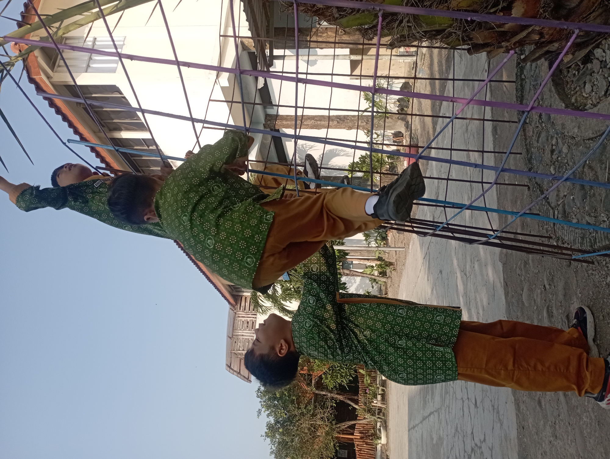 Two young boys are climbing a metal structure painted in various colors, including blue and purple, in what appears to be an outdoor schoolyard. The boy in the foreground is looking up and reaching for the structure. Both boys are wearing green patterned shirts with white designs and brown pants, suggesting a school uniform. Behind them, a white building with a red-tiled roof and multiple windows is visible. The ground is a mix of concrete paving and dirt. Green trees and foliage are present in the background. The sky is clear and blue. The overall scene suggests a warm, sunny day. The location is identified as Kramat, Indonesia.