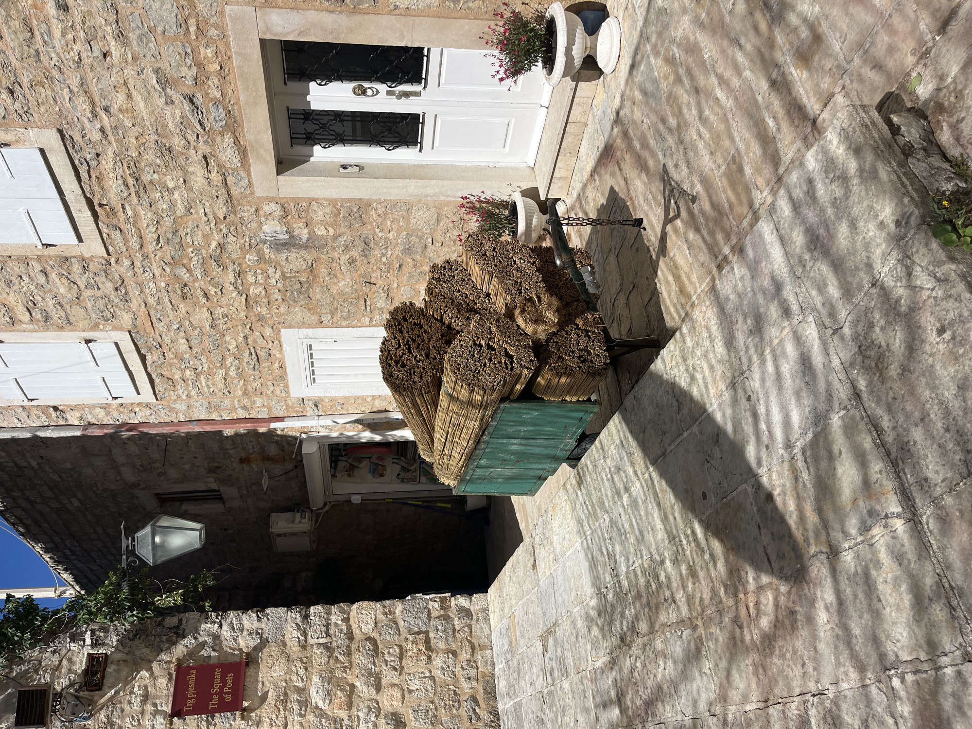 The image shows a narrow street scene in Budva, Montenegro. A stack of bundles of what appears to be reeds or straw rests on a teal-colored pushcart. The street is paved with stone blocks. The exterior of a stone building is visible on the left with white window shutters and a doorway with a white door and decorative ironwork. Above the door are two flowerpots filled with blooming plants. A sign reads "The Square of Poets." A hanging lantern is visible under an arched stone entryway. The light is bright, creating strong shadows on the stone pavement. There are no people visible in the image.