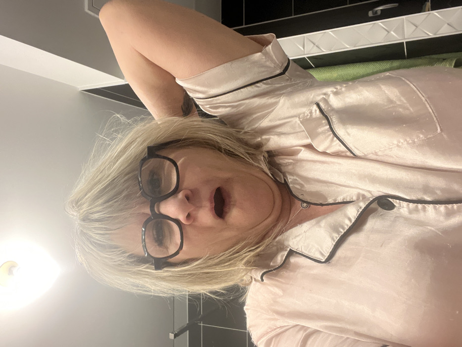 A woman in a pink, silky pajama top with black trim, wearing black-framed glasses, lies with her arm raised behind her head. She appears surprised, her mouth agape. Her blonde, fringed hair frames her face. The background includes bathroom elements like black tiling and a sink, with a light fixture visible to the left. A green towel hangs partially visible behind her.