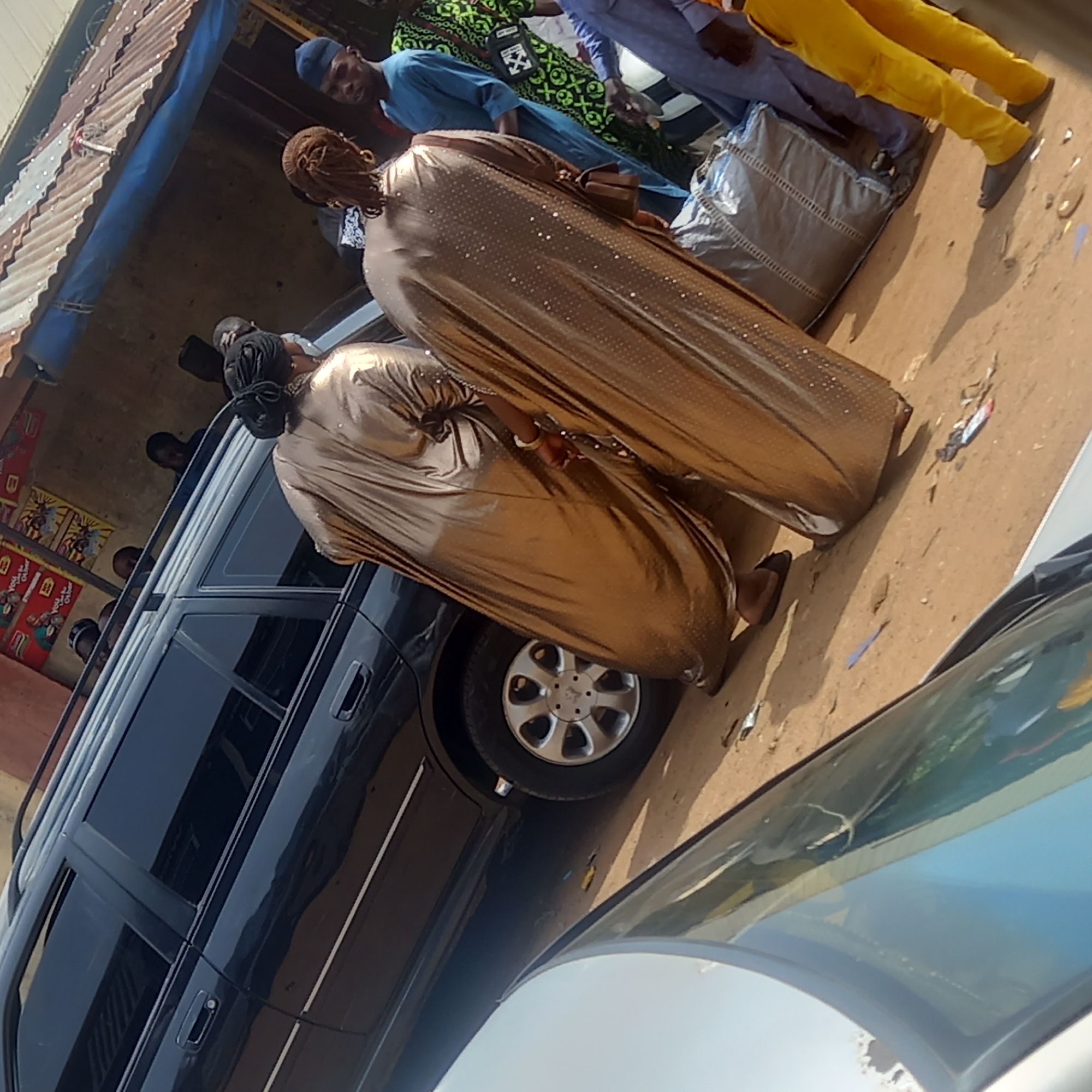 The image depicts a street scene in what is stated to be Dadi Riba, Nigeria. Two women in long, shimmering gold garments stand next to a black SUV. Other individuals are present in the background, including men in various clothing styles. A building with a corrugated metal roof is visible on the left. The ground appears to be dirt or sand. The view is partially obstructed by the windshield of another vehicle in the foreground.