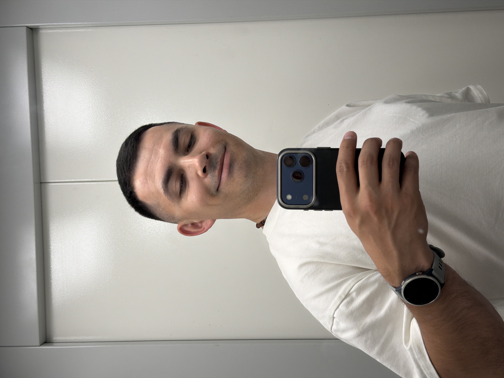 A slightly off-center selfie captures a man with short dark hair, light-medium skin, and stubble, appearing to be in his 20s or 30s. He is wearing a plain white short-sleeved t-shirt and a small dark necklace. On his left wrist, he sports a round-faced smartwatch with a grey and blue band. He holds a dark-cased smartphone, featuring prominent triple cameras, in his right hand, suggesting he is taking the photo in a mirror. He sports a relaxed smile with partially closed eyes, indicating contentment or amusement.
The background is a minimalist interior with plain light-colored, textured walls. Distinct vertical and horizontal lines suggest architectural features like wall panels or a door frame. Uniform indoor lighting illuminates the scene, making the time of day and weather indeterminate. There is no visible text within the image. While the location context is Izhevsk, Russia, the image itself provides no specific visual cues to identify the city.