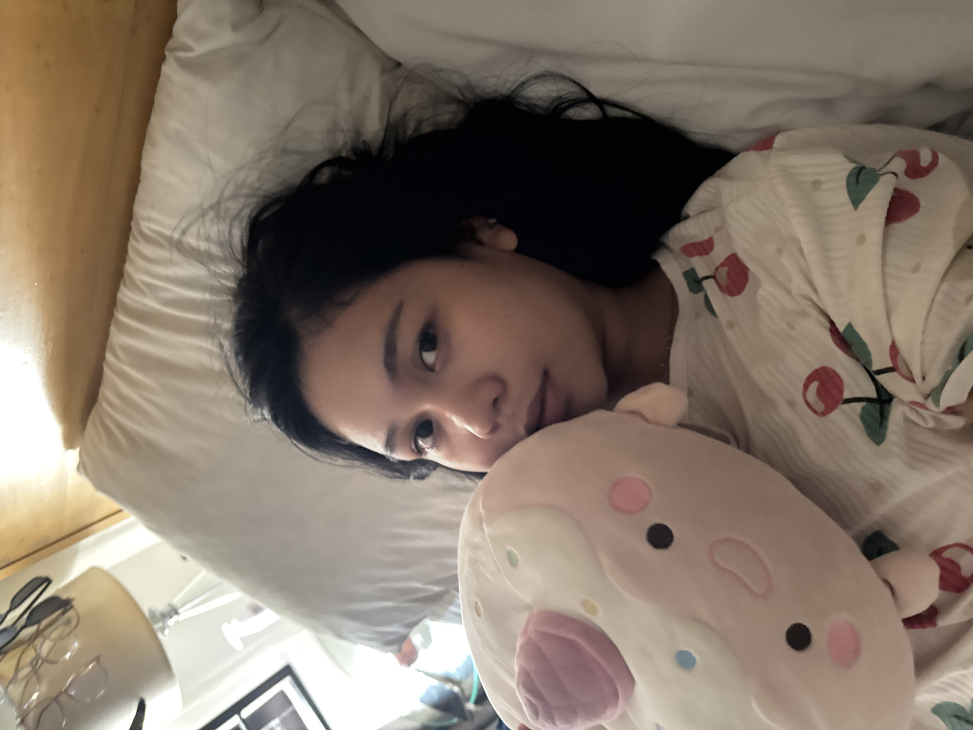 The image depicts an individual lying in a bed within an indoor setting. The person is a female, appearing to be a young adult of East Asian or Southeast Asian ethnicity, with long, dark hair spread across a white pillow. She is wearing a light-colored, ribbed top adorned with a red cherry pattern. Her gaze is directed towards the camera with a neutral expression.
She is holding a soft, plush toy against her chest. The toy is predominantly light pink and white, resembling a stylized dessert item like a donut or bun. It features two black eyes, two pink cheek spots, a pink oval-shaped mouth, a prominent rounded pink segment on its top, and several scattered multi-colored dots (light blue, light green, yellow, pink) suggesting sprinkles.
The bed is furnished with a white pillow and a white duvet or comforter. In the background to the left, a bedside lamp with a beige fabric shade is visible, on which multiple pairs of glasses or sunglasses are hanging. A black-framed picture is partially visible on the wall further to the left. The headboard of the bed is constructed from light-colored wood. The overall lighting suggests an interior room, likely a bedroom. No discernible visual cues within the image allow for the identification of a specific geographical location, including Davao City, Philippines.