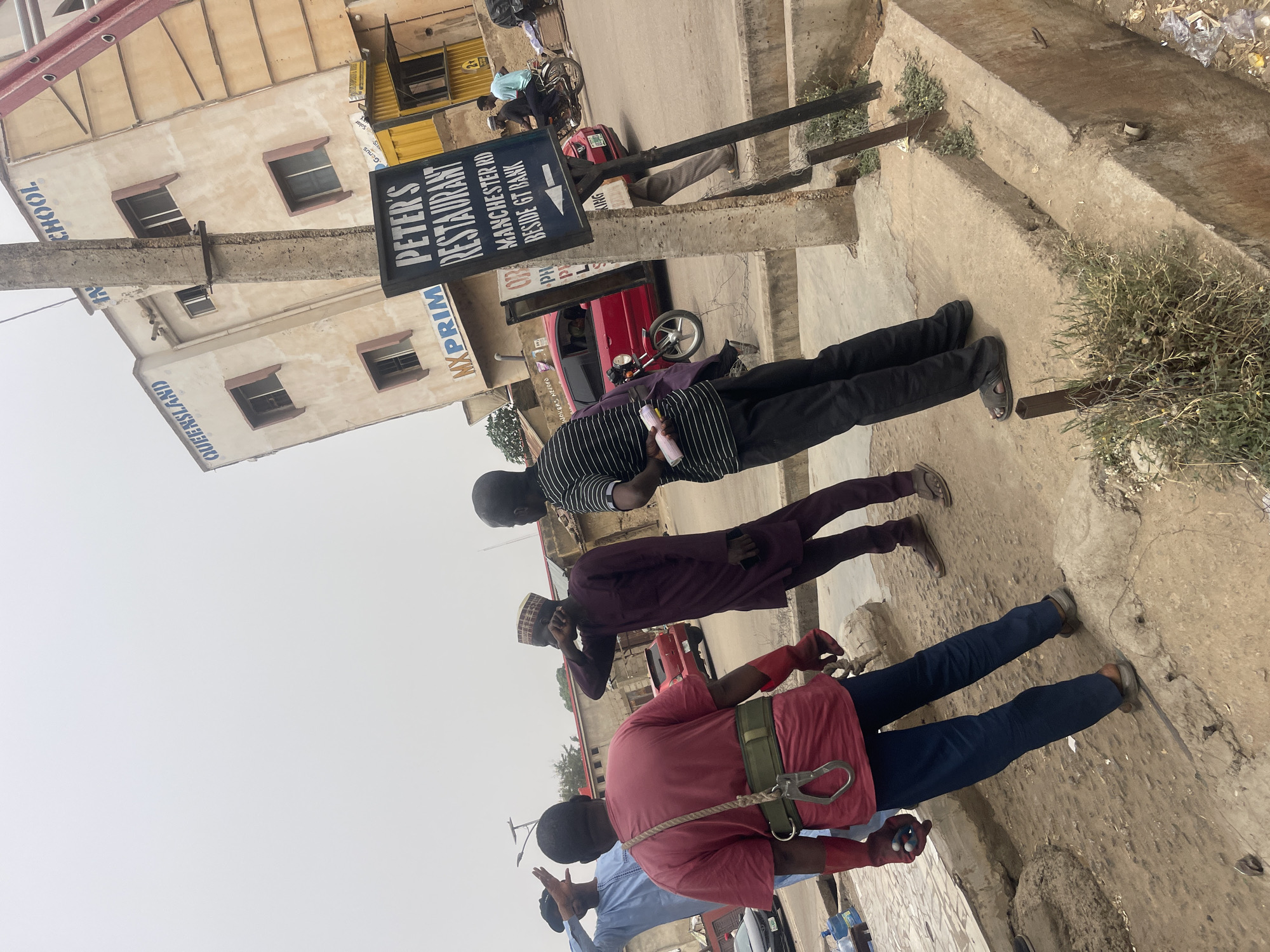 An urban street scene in Zaria, Nigeria, is depicted under an overcast or hazy daytime sky. The image primarily features several men along a concrete sidewalk next to buildings. In the foreground, a man wears a red shirt, blue pants, and red gloves, equipped with a harness and a carabiner. Beside him, another man in maroon traditional attire and a matching cap is focused on his mobile phone. Further into the midground, a man in a black and white striped shirt holds a small white object, possibly a bottle. Other individuals, a red car, a motorcycle, and a bicycle are visible in the background, suggesting general street activity.
Light-colored multi-story buildings line the street. One building clearly shows "QUEENSLAND SCHOOL" painted on its facade. A prominent blue sign, attached to a concrete pole, advertises "PETER'S RESTAURANT" with an arrow pointing right. Additional text on this sign includes "WORKERS NEEDED," "MXPRIM," "MANCHESTER RD," "PH," and "BESIDE GT BANK," along with "Genus" and "Solar." A smaller, faint sign reads "KING." The ground consists of concrete, with patches of green vegetation growing from cracks and edges. A red ladder is partially visible leaning against a building in the distance. The scene captures a slice of daily life in a bustling urban environment.